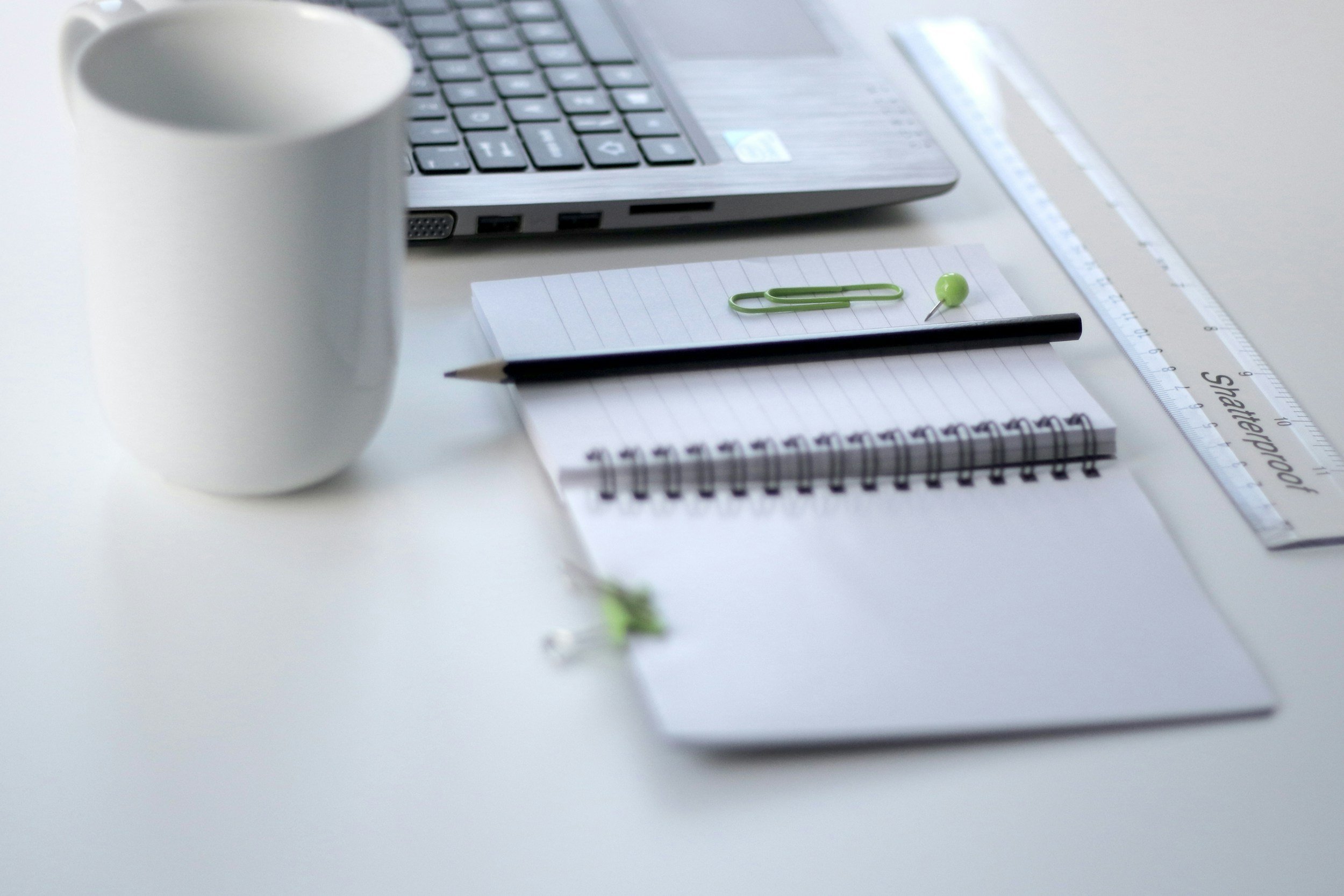 A white desk with a white mug, a laptop, a black pen, a spiral-bound notebook, a green paper clip, a green push pin, and a clear ruler.