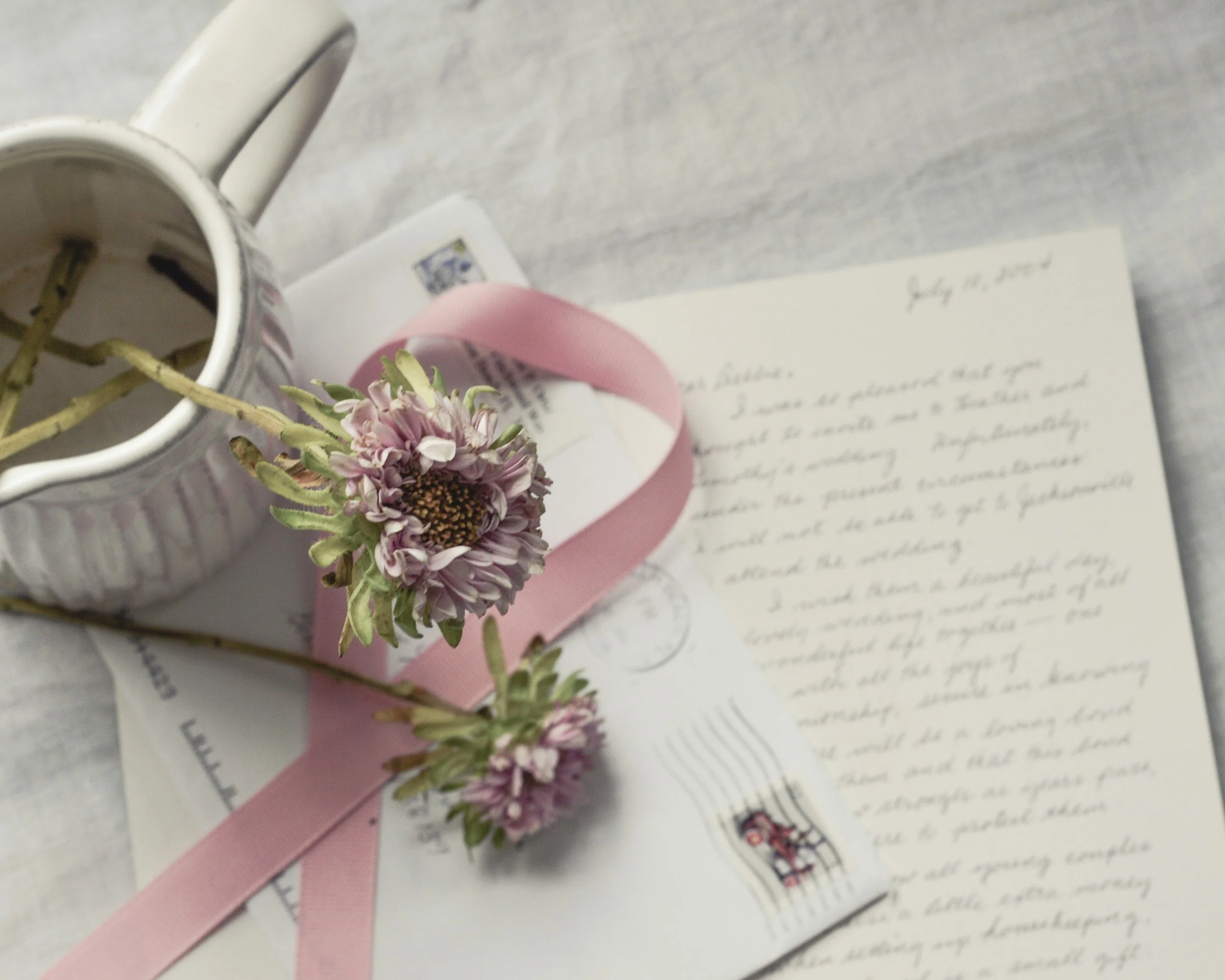 A pink flower with green leaves resting on a letter, a pink ribbon, and a white mug with a small branch inside, on a light-colored surface.