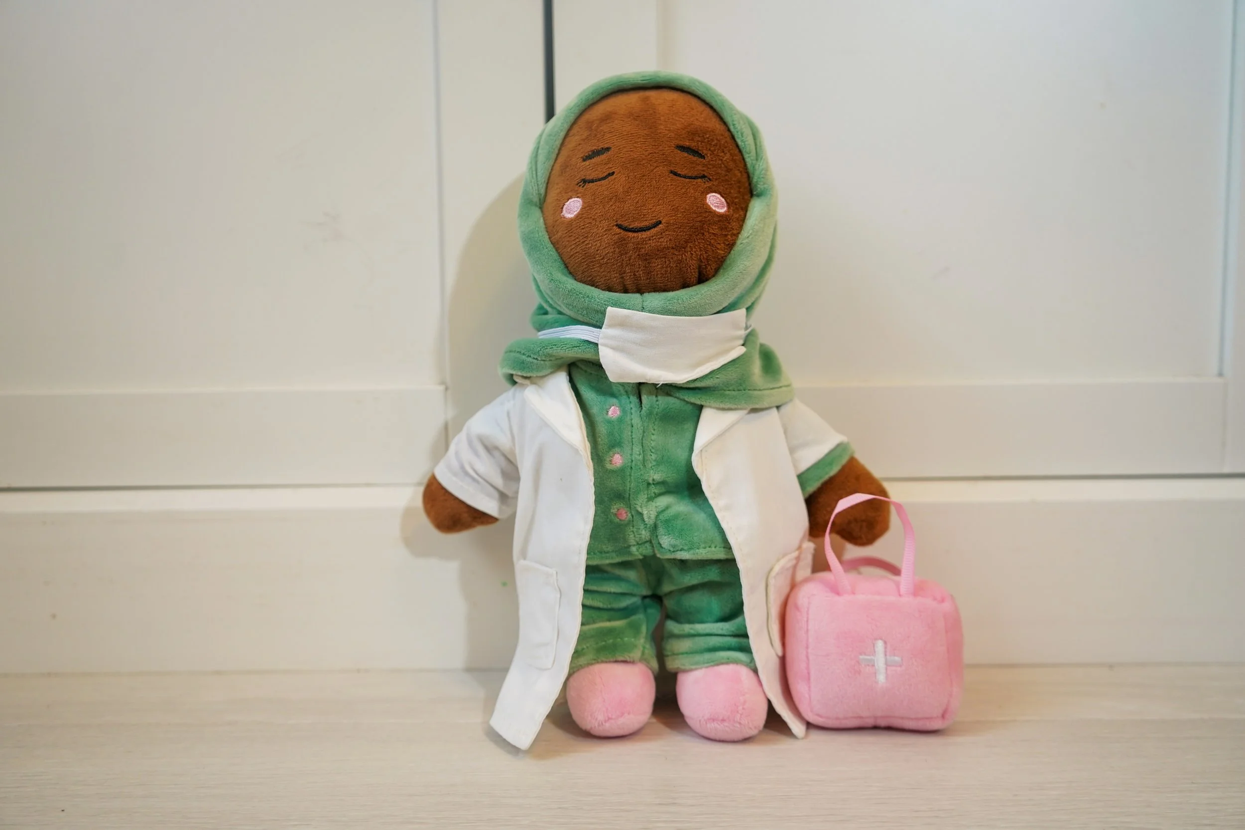A plush muslim female doll dressed as a healthcare worker, with a brown face, pink cheeks, and closed eyes, wearing a green hoodie, white coat, green scrubs, pink slippers, and holding a pink first aid kit.