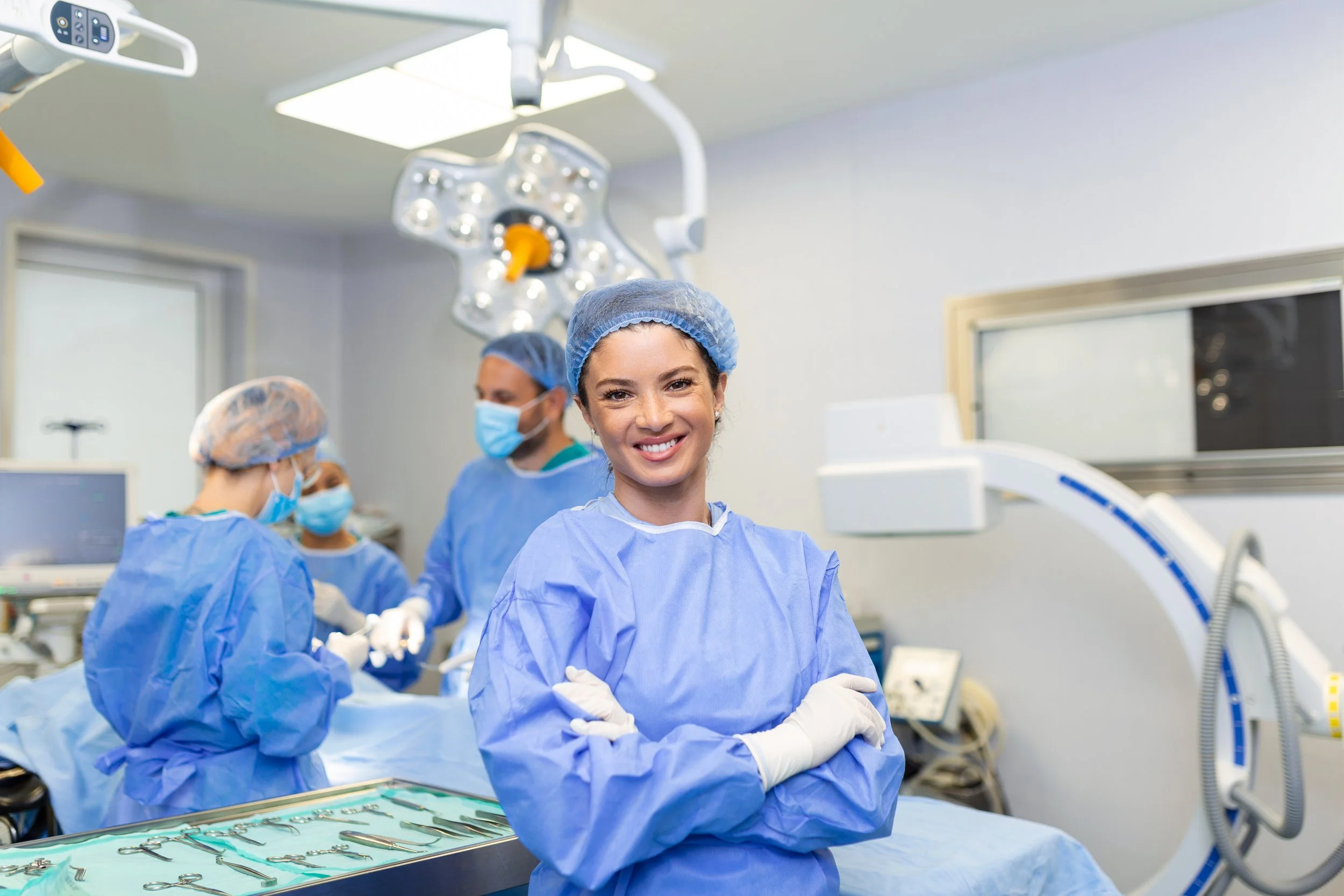 A smiling female surgeon in scrubs and a hair cap standing with arms crossed in an operating room with surgical staff and equipment in the background.