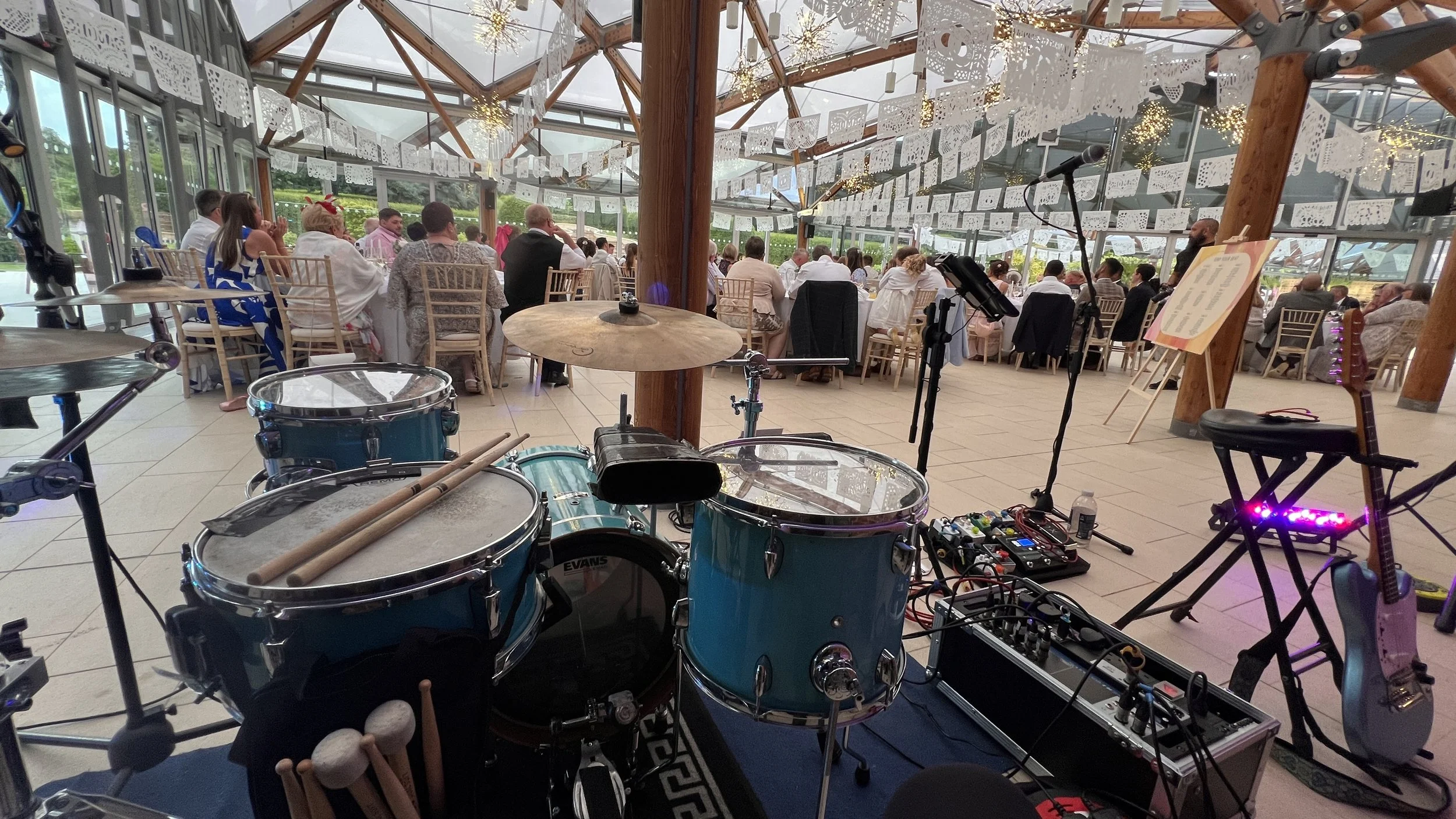 Stage setup with a drum set, microphone stand, electric guitar, music pedals, and other audio equipment, overlooking a large banquet hall filled with people seated at tables, decorated with white paper cutouts and string lights.