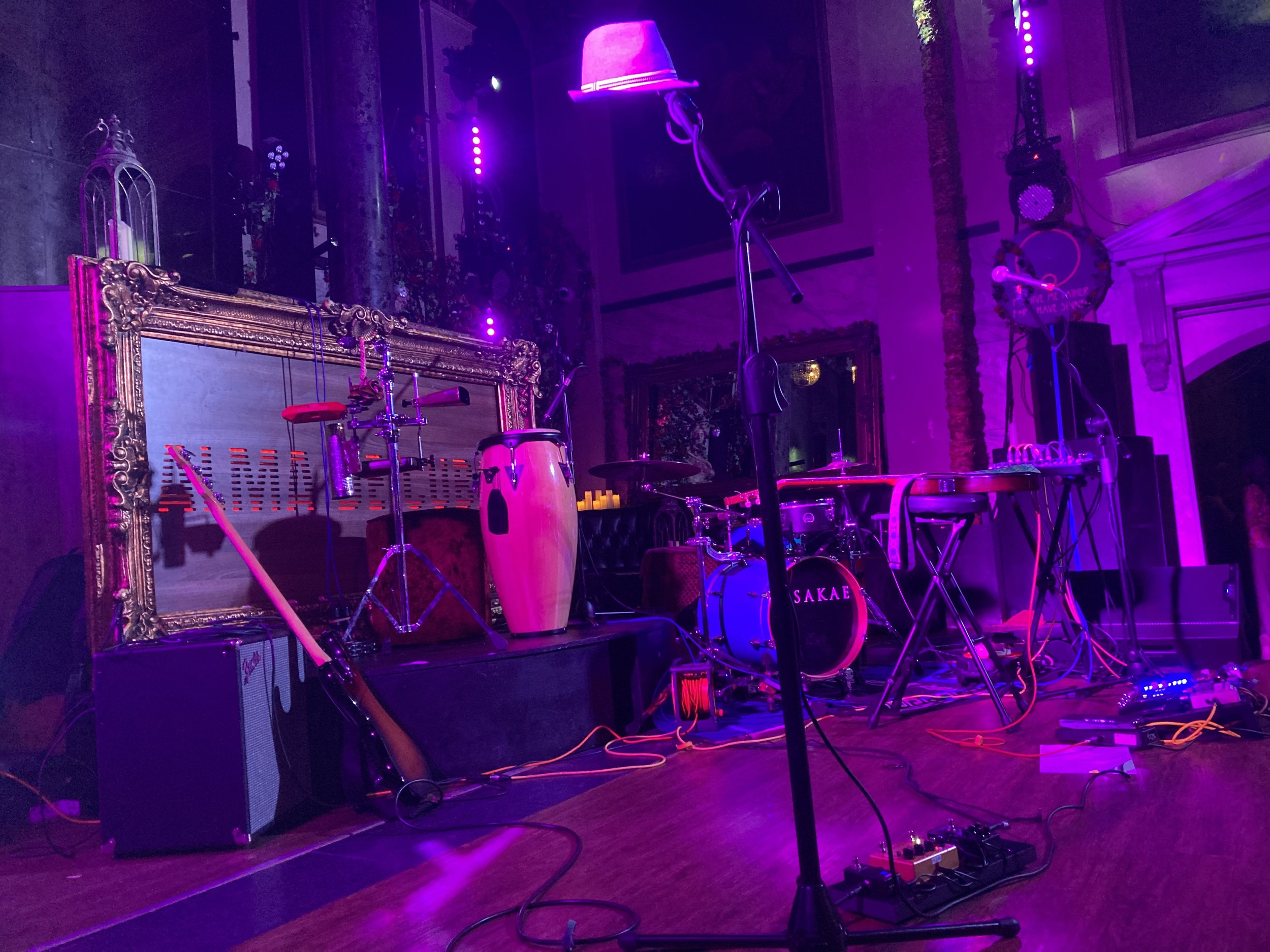 Stage setup with musical instruments including drums, a guitar, conga, keyboard, and microphones, illuminated by purple lighting in an indoor venue.