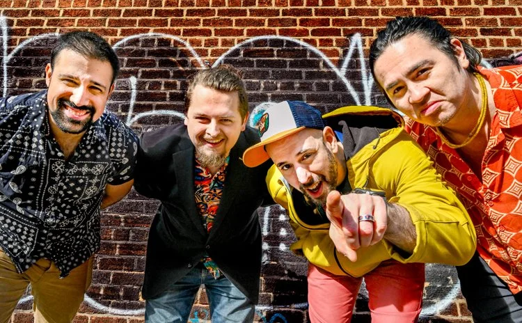 Four men smiling and posing in front of a brick wall with graffiti, one pointing at the camera. Guacamaya Latin Band also creates original music. Their album will be out soon.