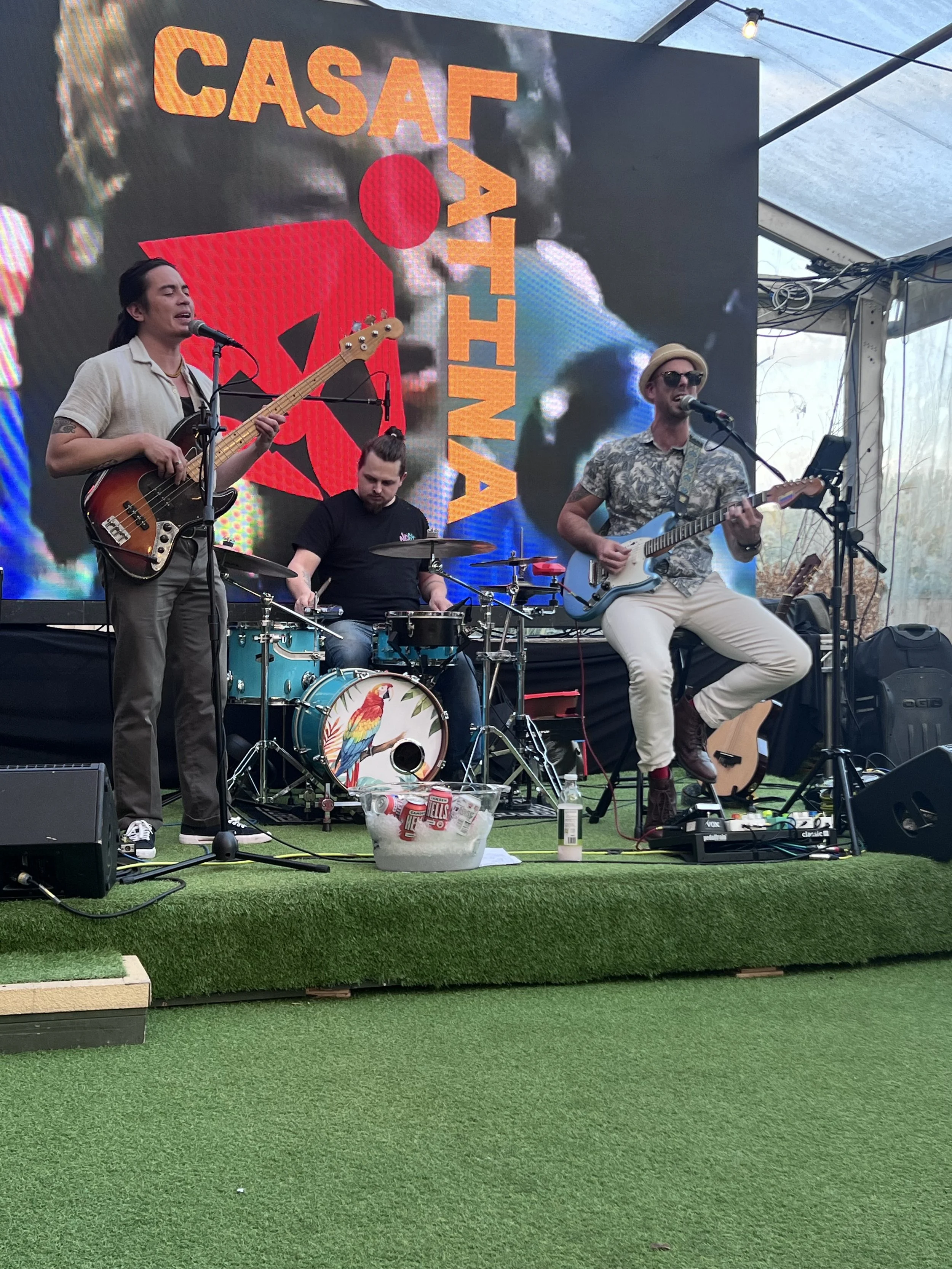 Three musicians performing on a stage with a large digital display behind them. The display shows the SASAF logo with a red dot and the word 'ATTINA' vertically. One musician is playing a bass guitar, another is on drums with a parrot design on the bass drum, and the third is singing and playing an electric guitar, wearing sunglasses and a hat. There are supplies and equipment on the stage, including a bucket of ice with drinks, a bottle, headphones, and speakers.