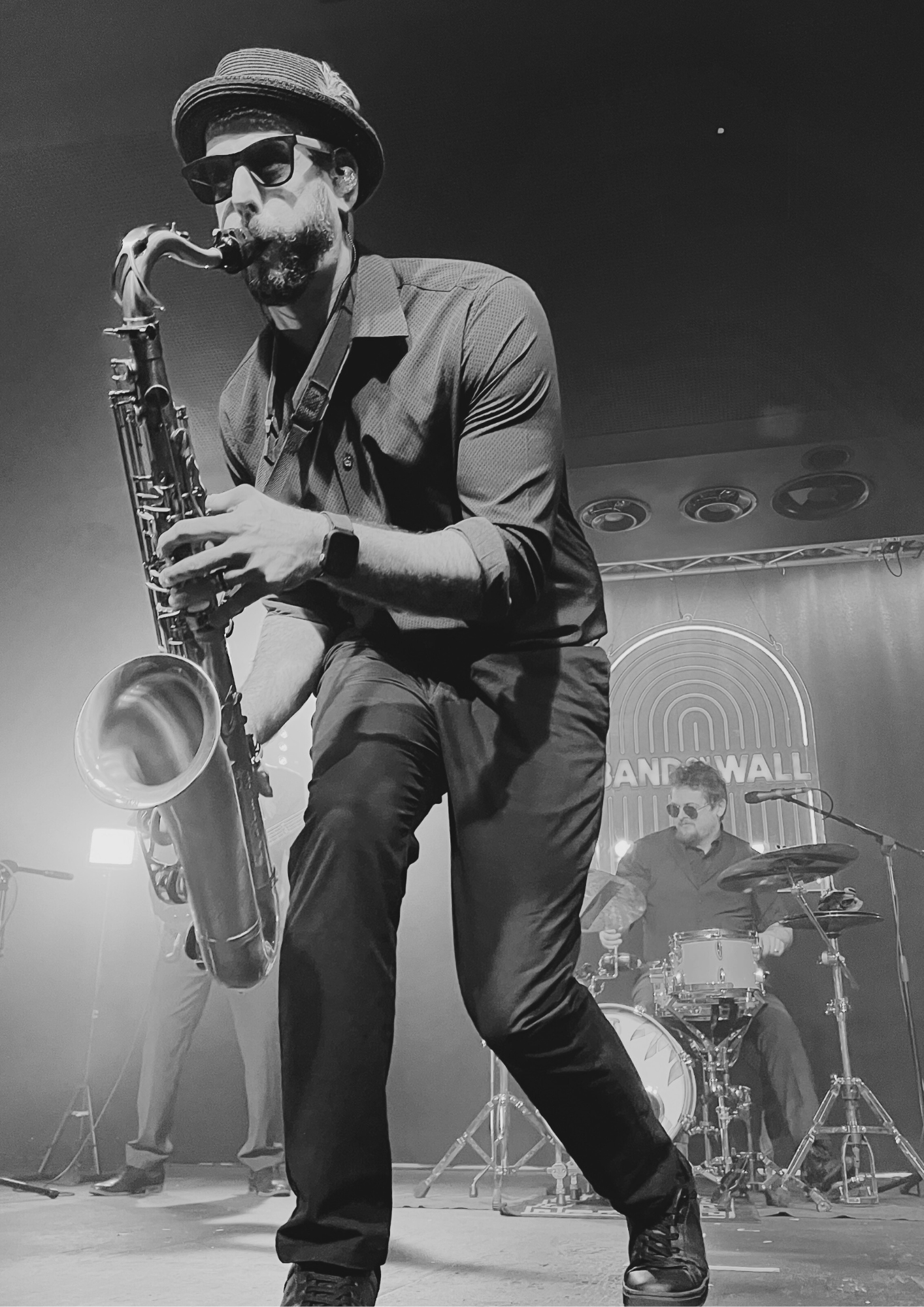 Best Latin Band in Manchester, UK. With the most positive reviews Latin band in the north west of the UK. Musician playing saxophone on stage with drummer in background, stage lit with professional lighting the words 'Band on the wall'.