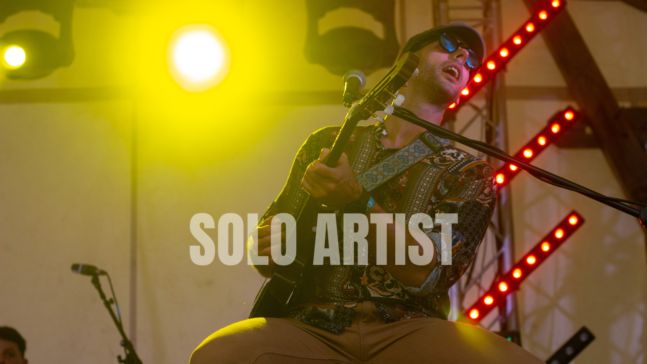 Solo artist who performs British Pop, Latin Music, African music, and a variety of world music styles and genres. The singer is wearing sunglasses, a patterned shirt, and a hat, playing an acoustic guitar on stage with colorful stage lights.