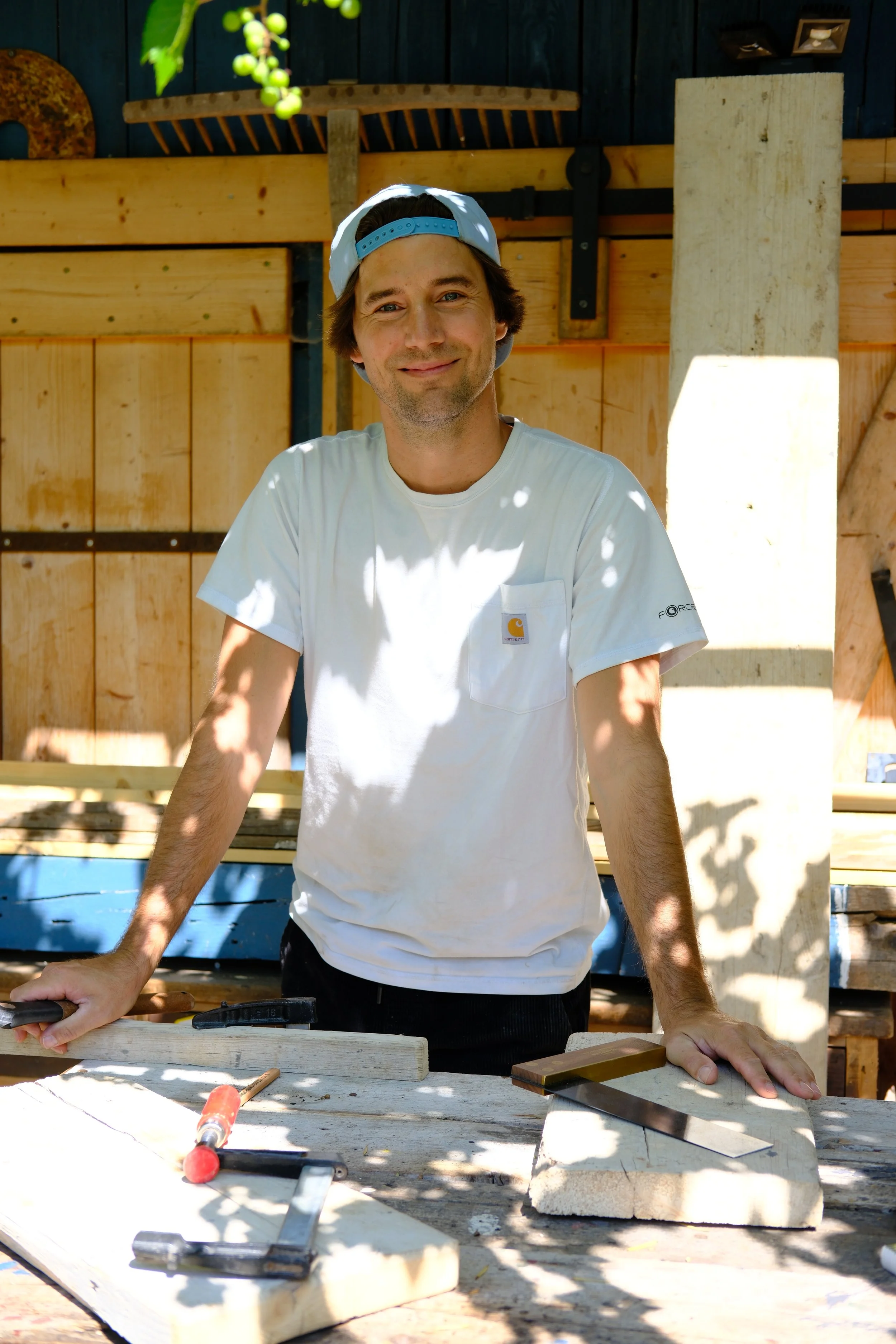 Moritz arbeitet in einem weißen T-Shirt und einer blauen Cap an einem Holzprojekt in einer Werkstatt. Er steht vor einer Werkbank mit Werkzeugen und Holzstücken.