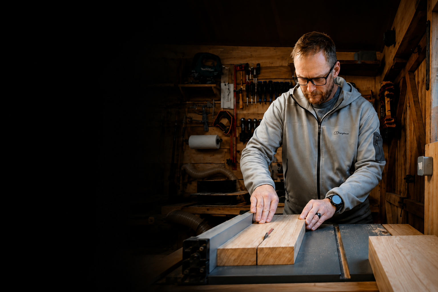 A man wearing safety glasses and a gray jacket working with wood in a woodworking shop, cutting a piece of wood on a table saw.