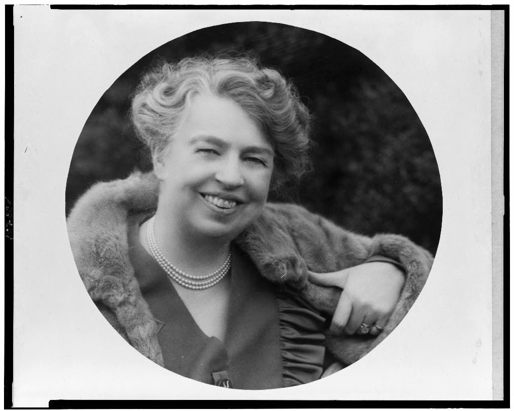 A black and white portrait of a smiling Eleanor Roosevelt with short, curly hair, wearing a pearl necklace, a dark dress, and a fur stole over her shoulders, with a dark blurred background.