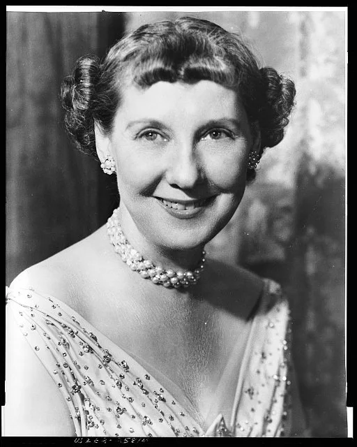 Black and white portrait of a smiling Mamie Doud Eisenhower with short, curled hair wearing a pearl necklace and pearl earrings, dressed in a beaded evening gown.
