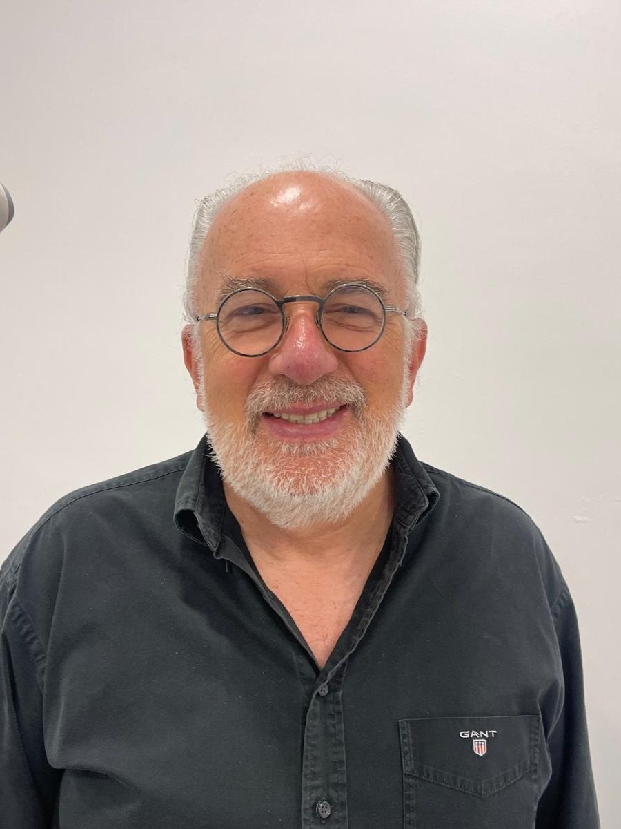 A middle-aged man with glasses and a white beard, smiling, wearing a black GANT shirt, standing against a plain white wall.