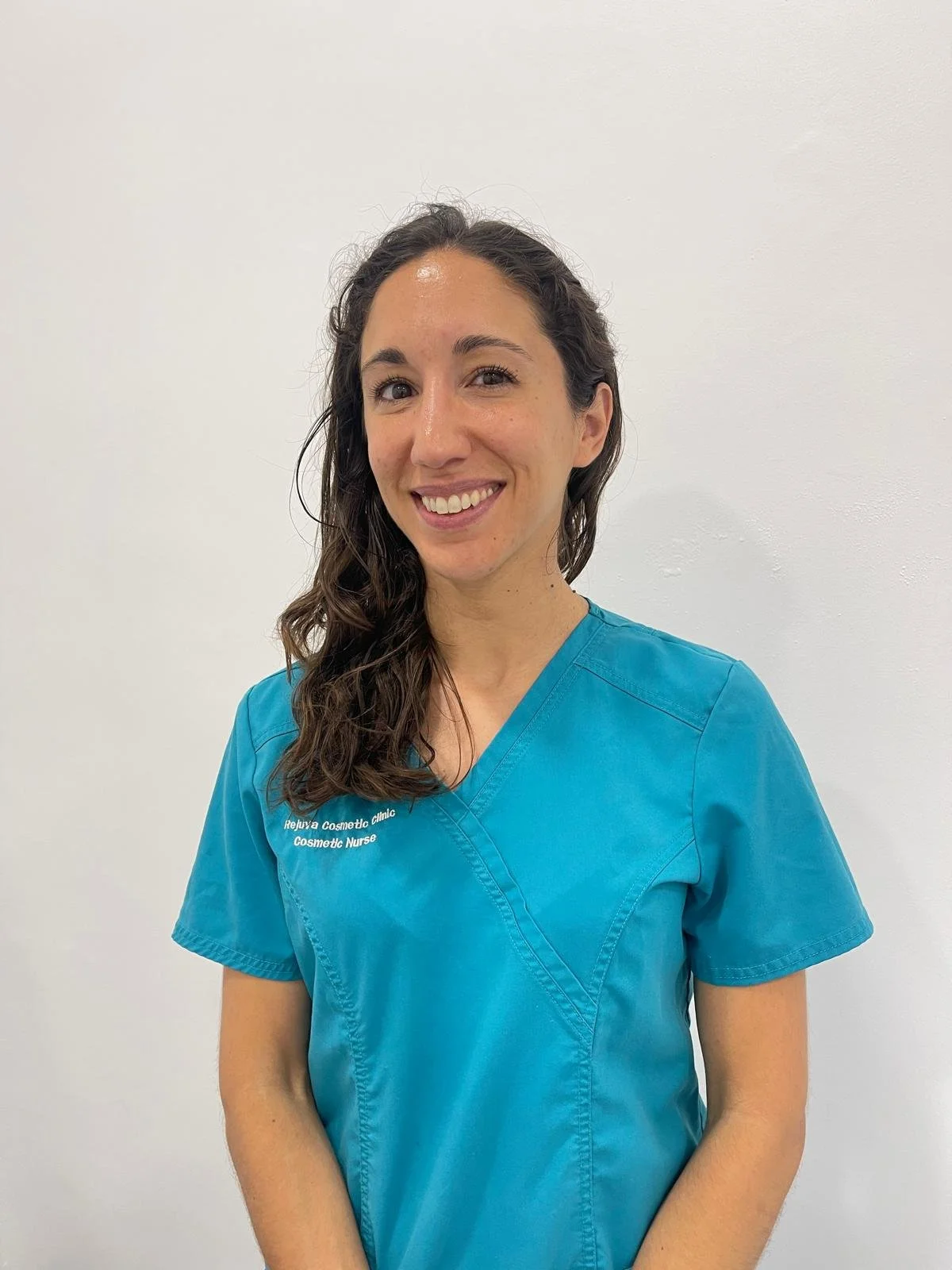 A woman wearing blue medical scrubs with a name tag that reads 'Rejuva Cosmeto Clinic, Cosmetic Nurse' stands against a plain white background, smiling at the camera.
