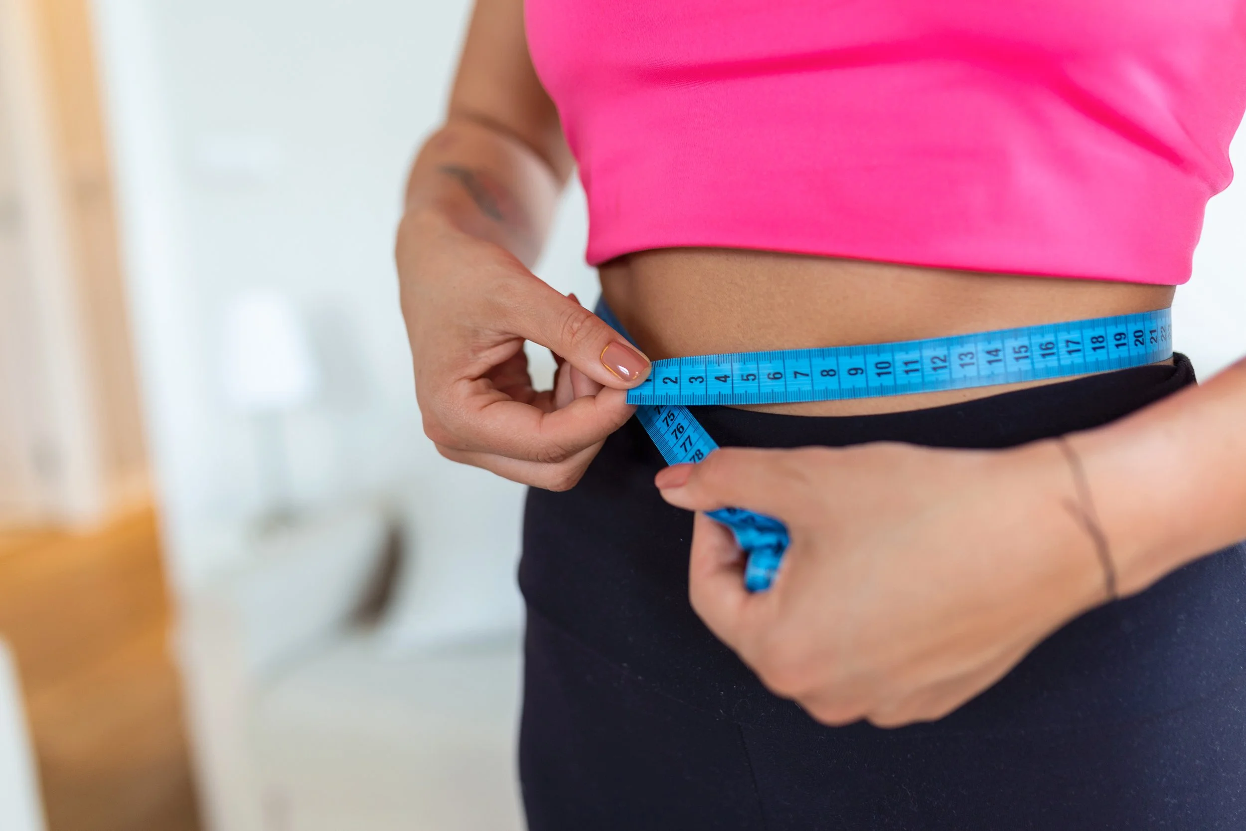 Person wearing a pink top measuring their waist with a blue tape measure.