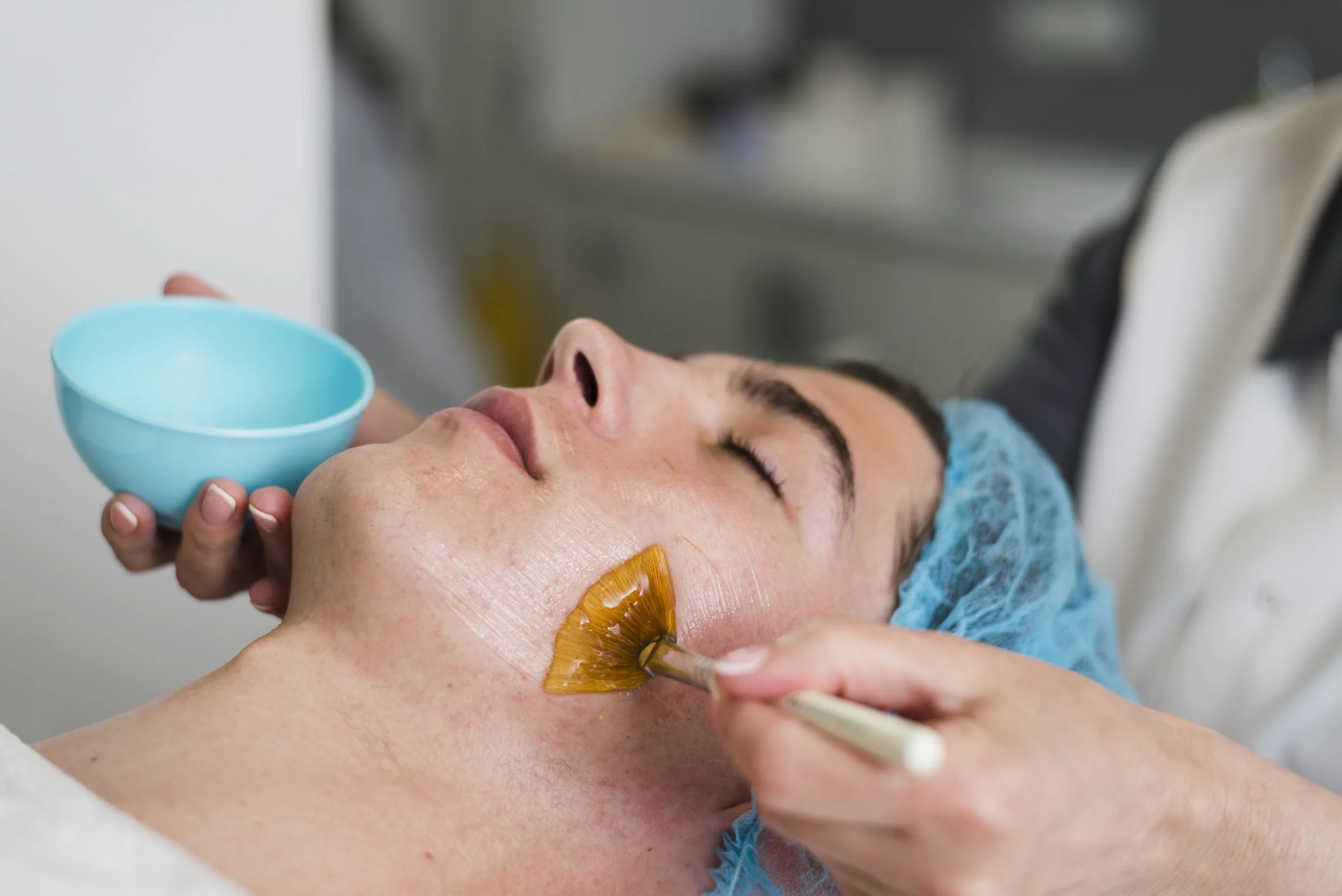 A person receiving a facial treatment, lying down with a hair cap, a brush applying a mask on their cheek, and holding a bowl.