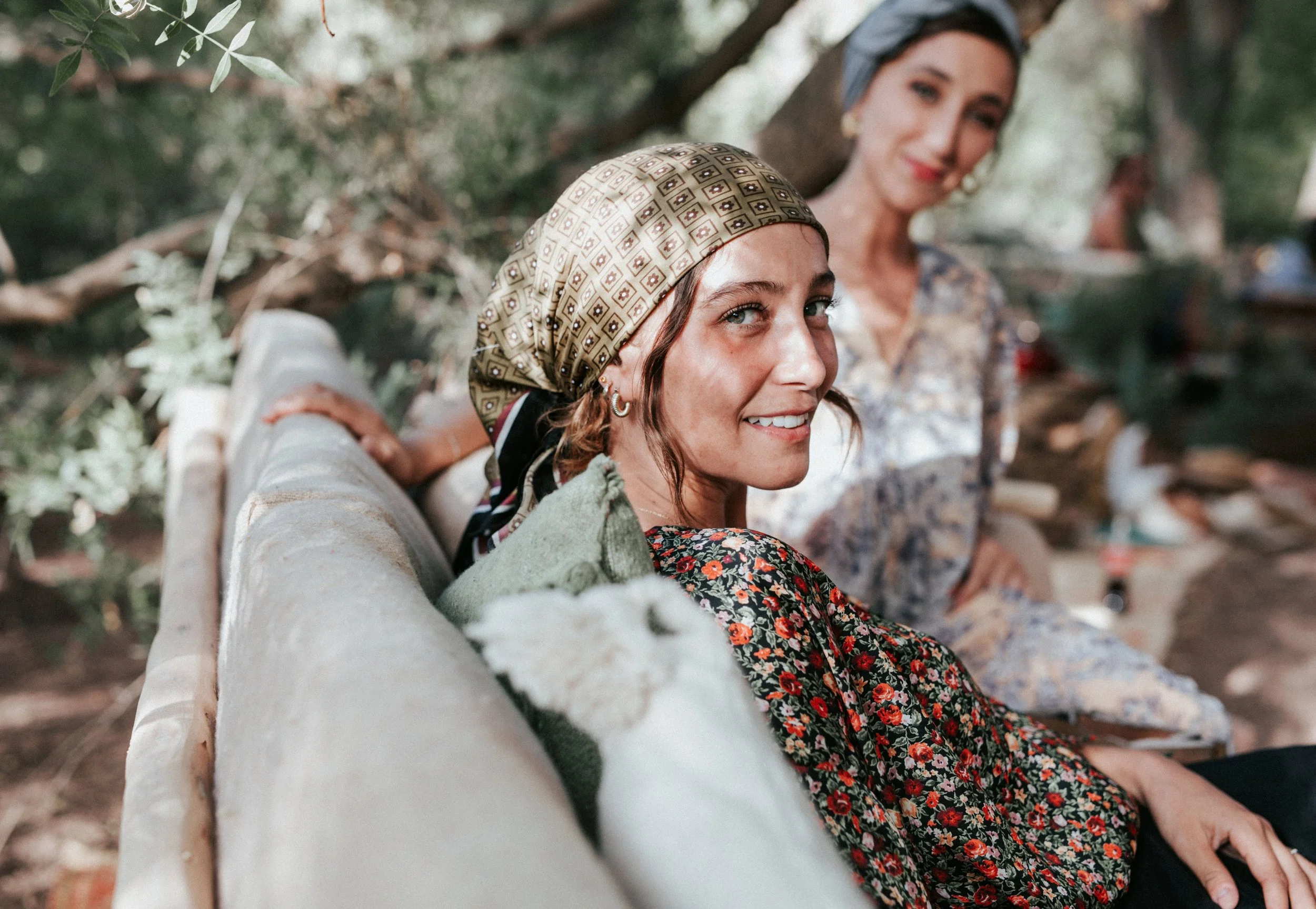 Deux femmes assises sur un banc en pleine nature, l'une porte un foulard beige avec un motif géométrique, l'autre porte un foulard gris et une robe à motifs floraux, elles sourient.