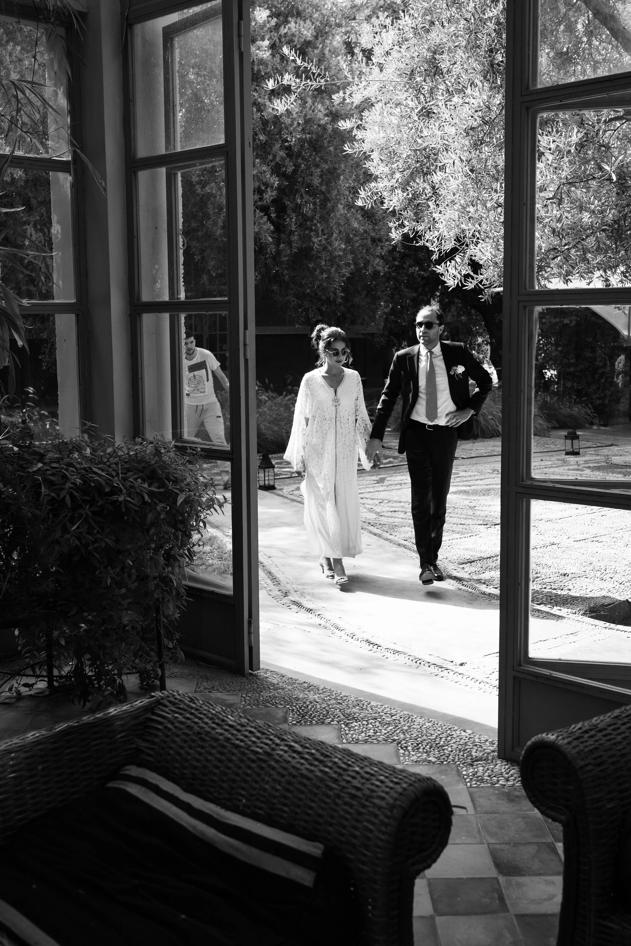 Un couple habillé formellement, marche main dans la main à l'extérieur, vue depuis une porte ouverte, avec un décor naturel et un homme en arrière-plan.