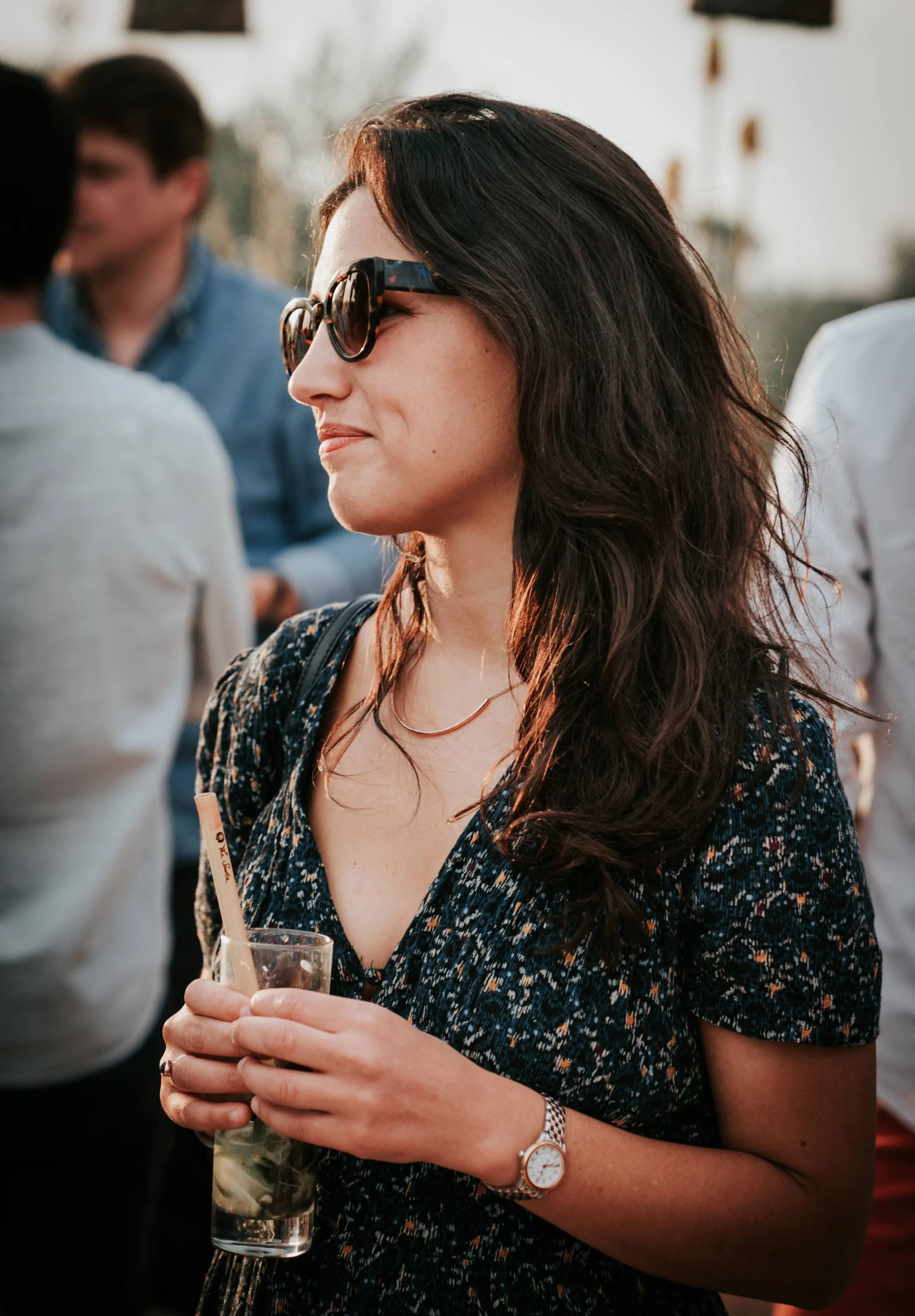 Une femme portant des lunettes de soleil noires, une robe noire à motifs, un bracelet en or, une montre en argent et un collier, tient un verre avec une paille lors d'un événement en plein air, entourée d'autres personnes.