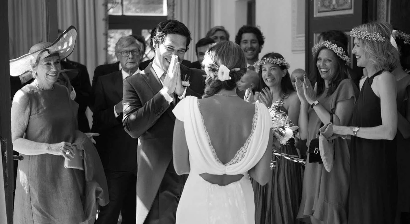 Un couple lors de leur mariage entouré d'invités, la femme porte une robe de mariage et des fleurs dans ses cheveux, le marié en costume, tout le monde sourit et semble heureux.