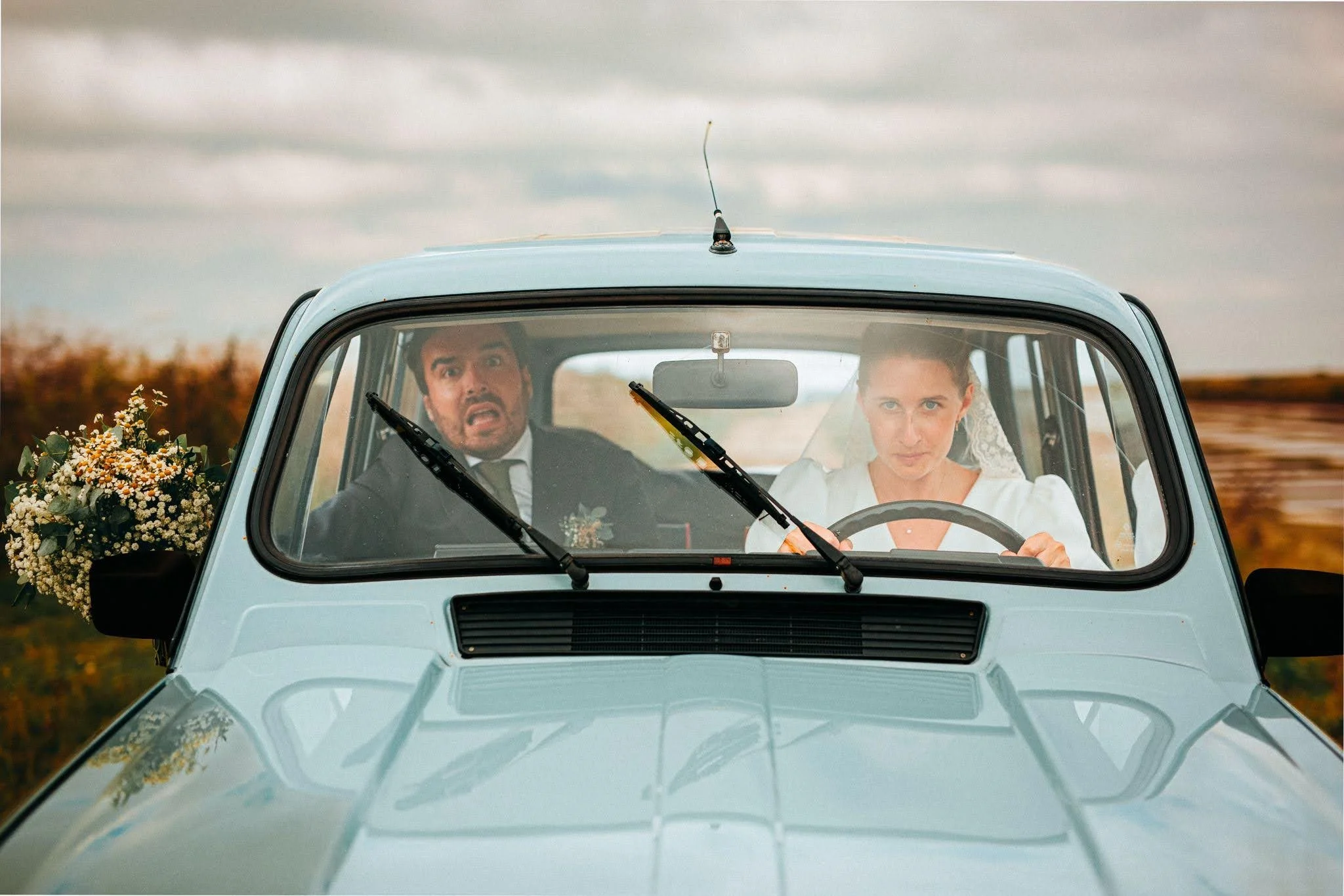 Un couple en voiture, probablement un mariage, avec un homme en costume et une femme en robe blanche, semblant nervieux ou surpris. La voiture est claire et il y a un bouquet de fleurs à l'arrière.