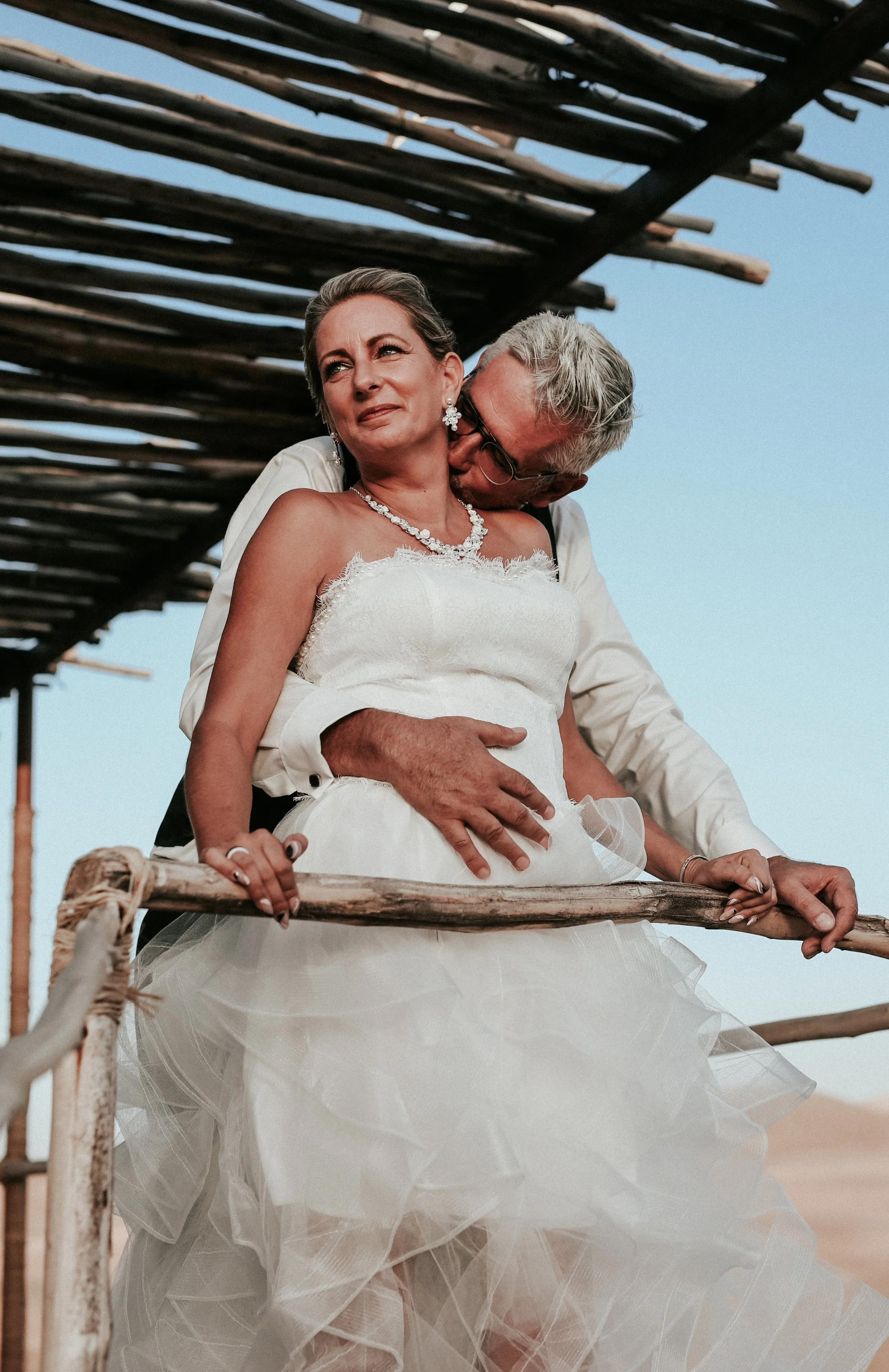 Un couple en mariage, la femme porte une robe blanche et des bijoux, l'homme l'embrasse tendrement dans leur dos, sous une structure en bois, en extérieur, avec un ciel bleu.