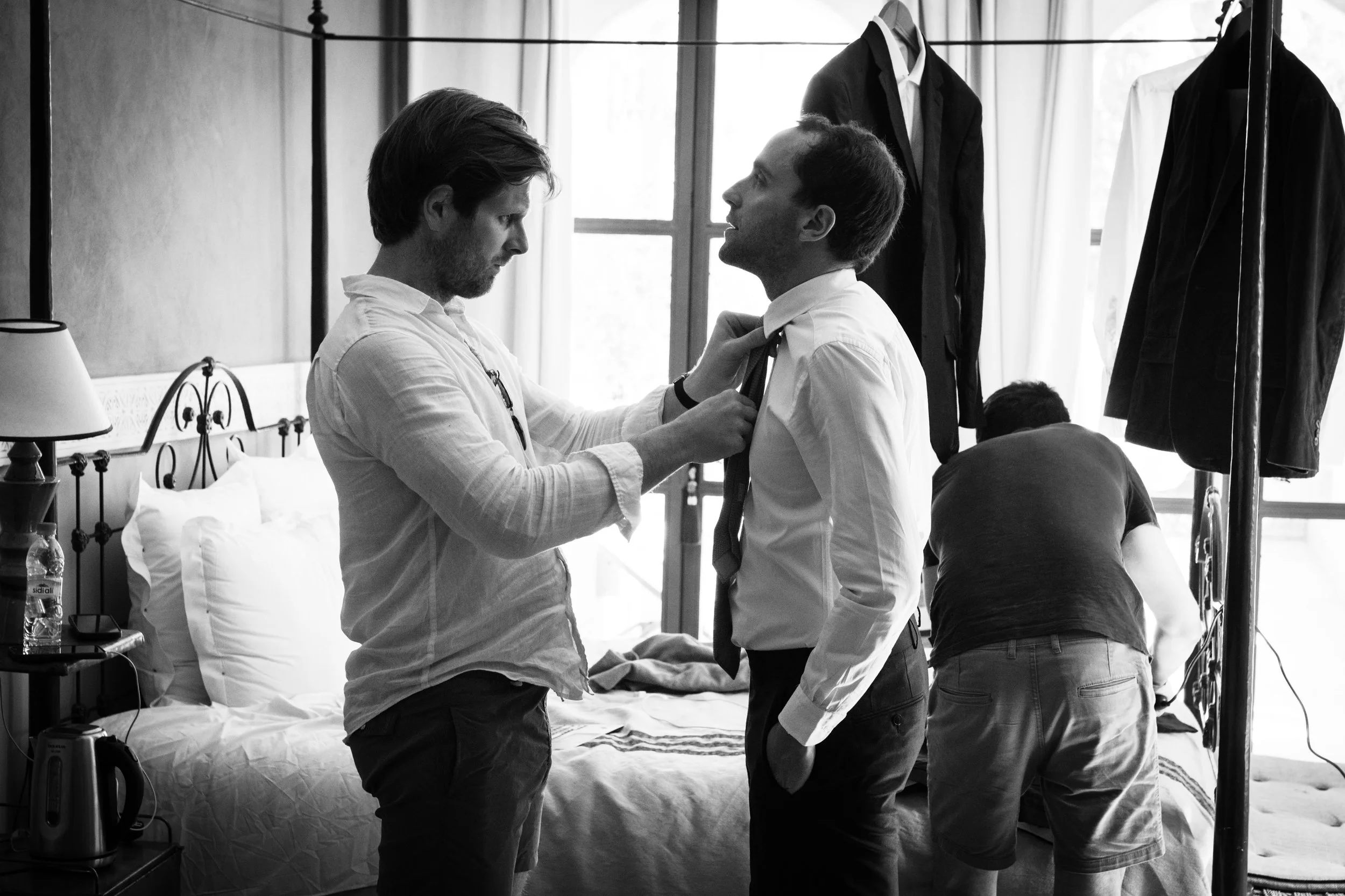 Deux hommes se préparent dans une chambre, l'un ajustant la cravate de l'autre, avec des vêtements suspendus en arrière-plan.