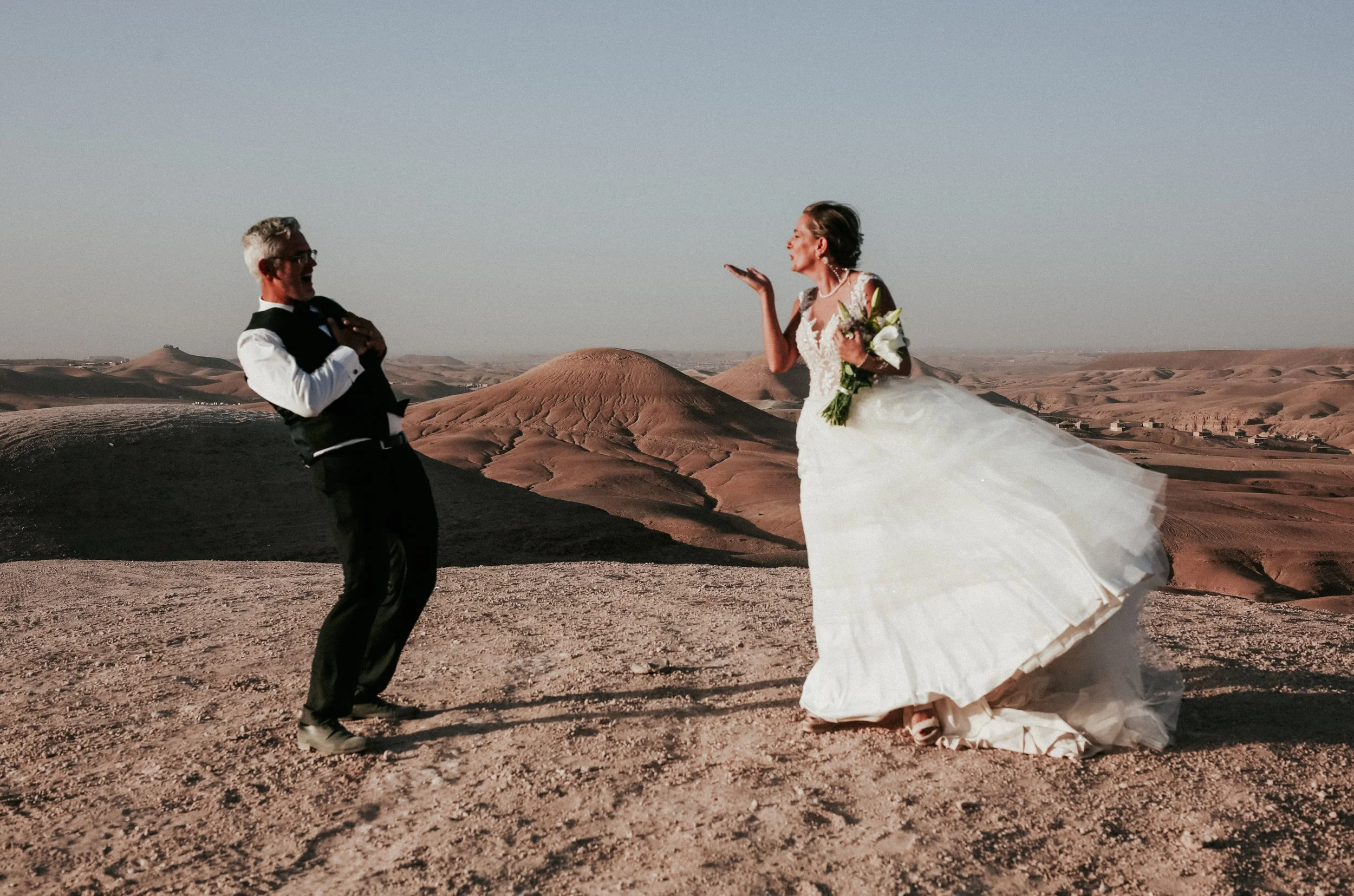 Un couple en mariage, une femme en robe blanche et un homme en costume, dans un désert avec des dunes en arrière-plan, avec une atmosphère joyeuse et romantique.