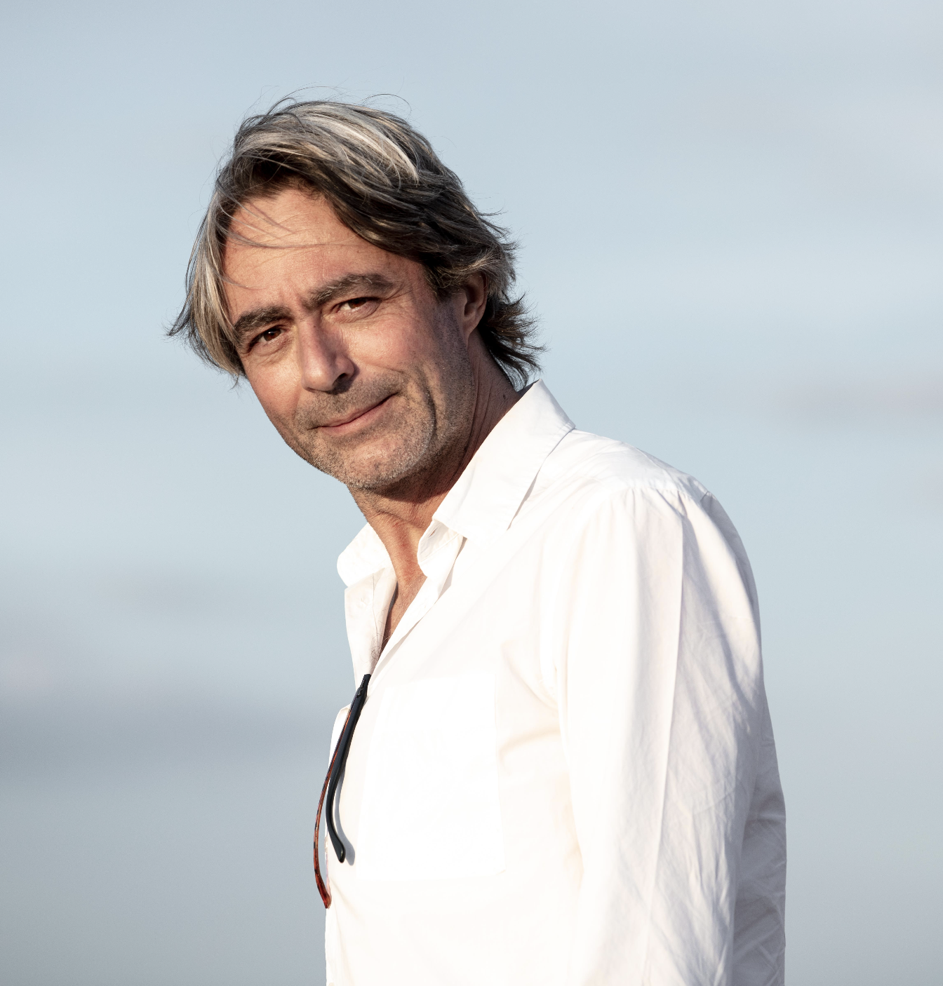 Un homme barbu, avec des cheveux gris, portant une chemise blanche, se tient contre un ciel bleu.