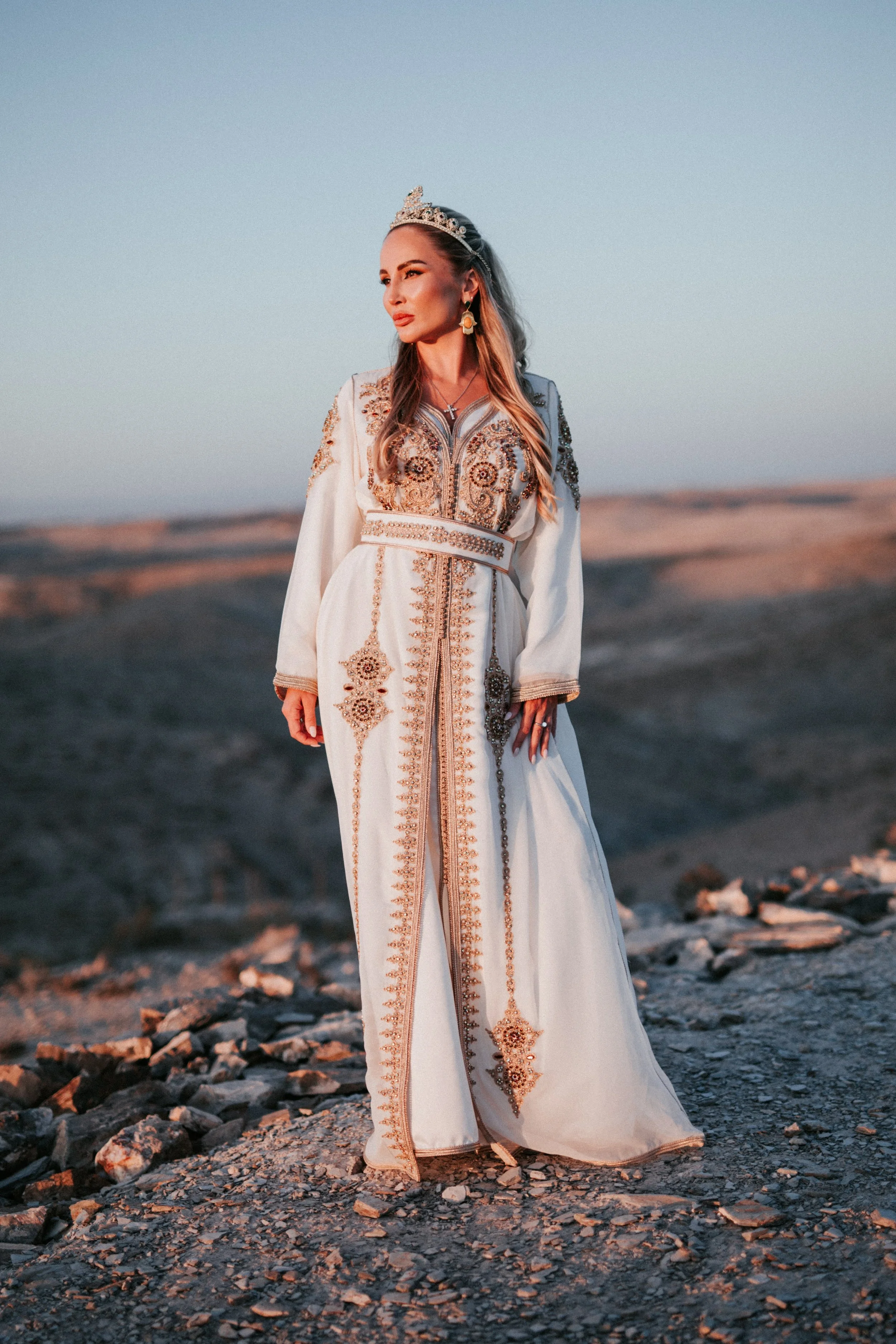 Femme habillée d'une robe blanche ornée de broderies dorées, portant une couronne et des boucles d'oreilles, dans un paysage désertique au coucher du soleil.