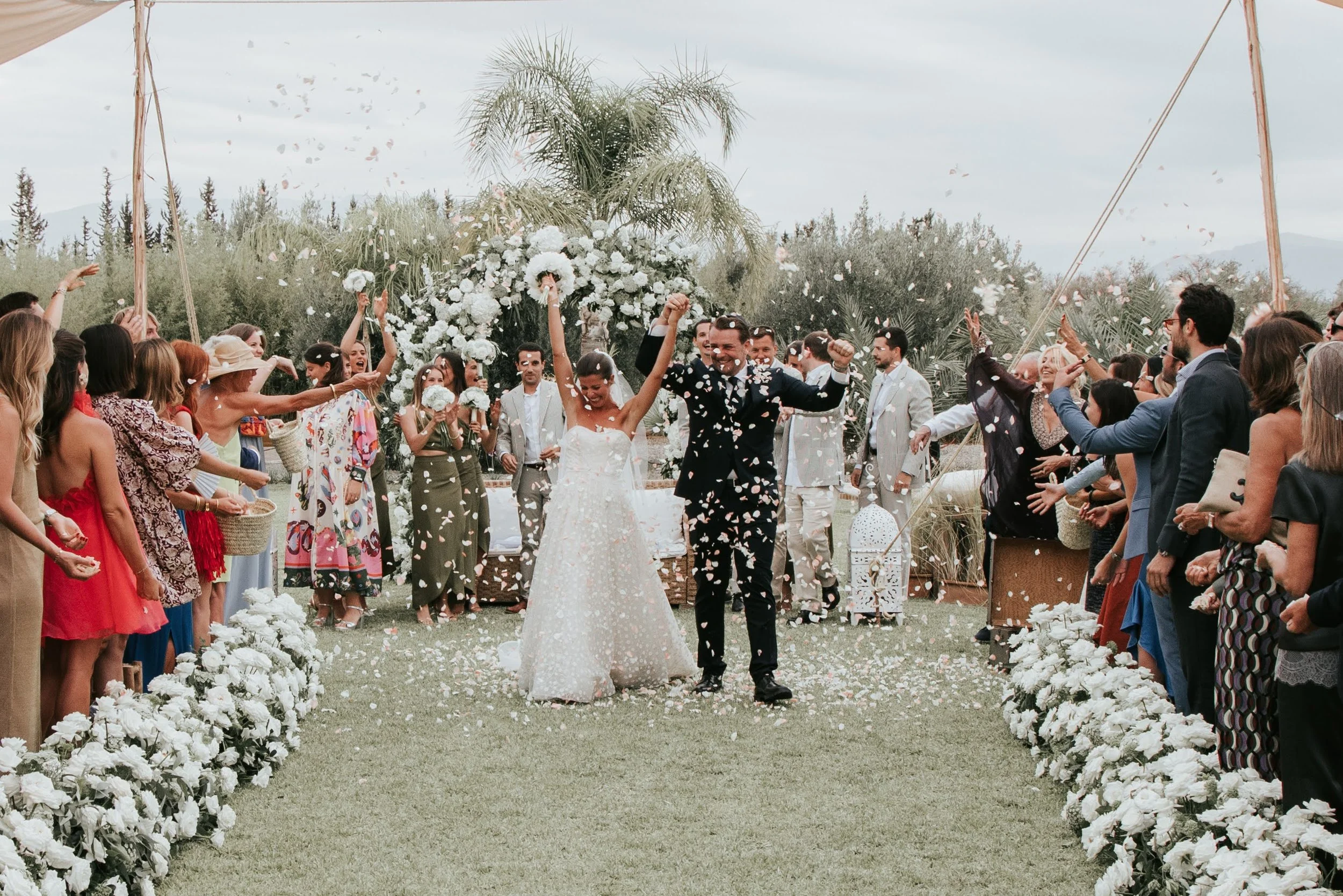 Un couple de mariés souriant et levant les bras, entouré d'invitée en train de célébrer lors d'une cérémonie de mariage en extérieur. La scène est décorée avec des fleurs blanches et un grand arbre en arrière-plan.