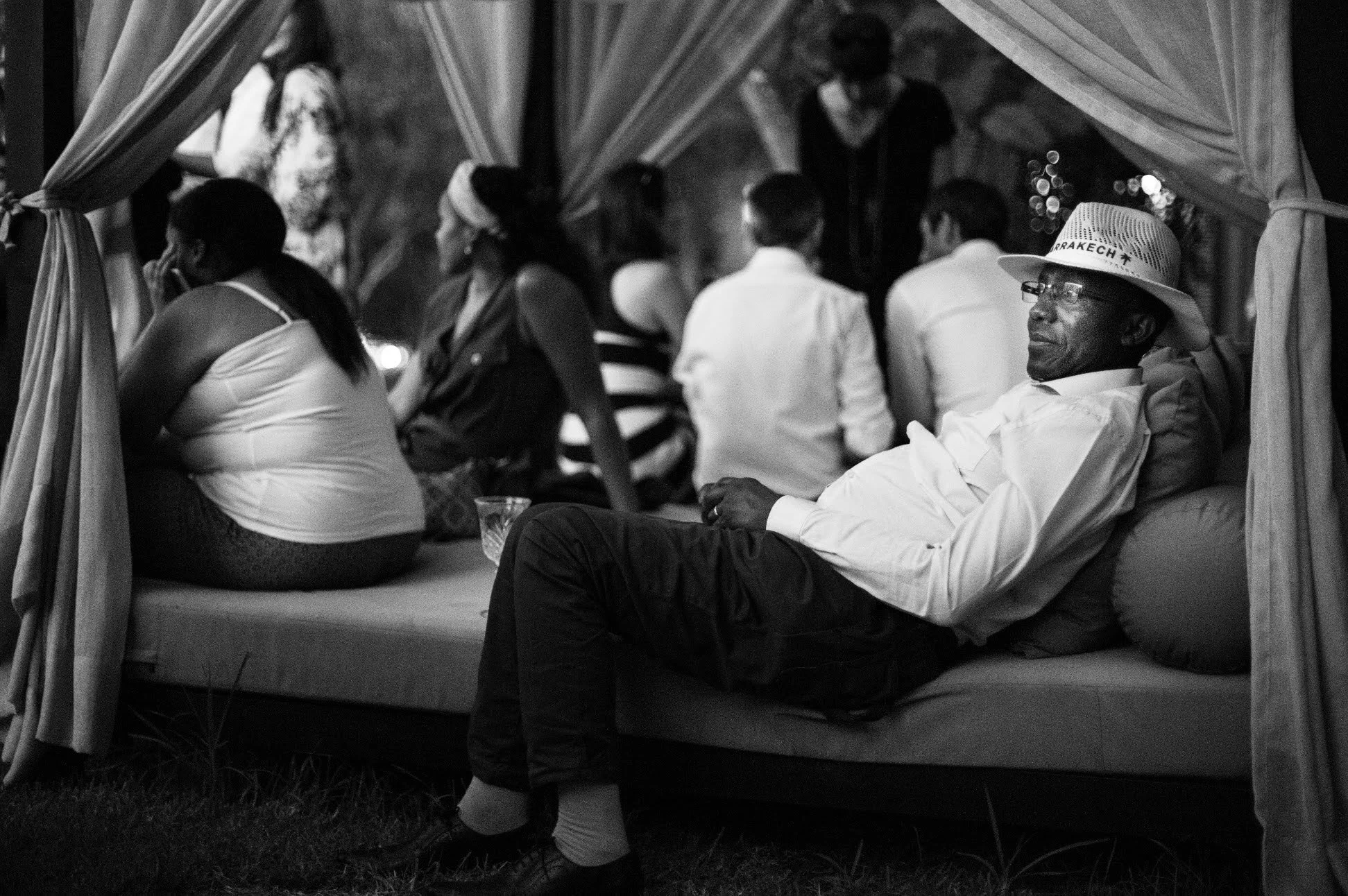 Un homme portant un chapeau et des lunettes, assis sur un canapé, avec d'autres personnes assises derrière lui dans ce qui semble être un événement ou une fête, en noir et blanc.