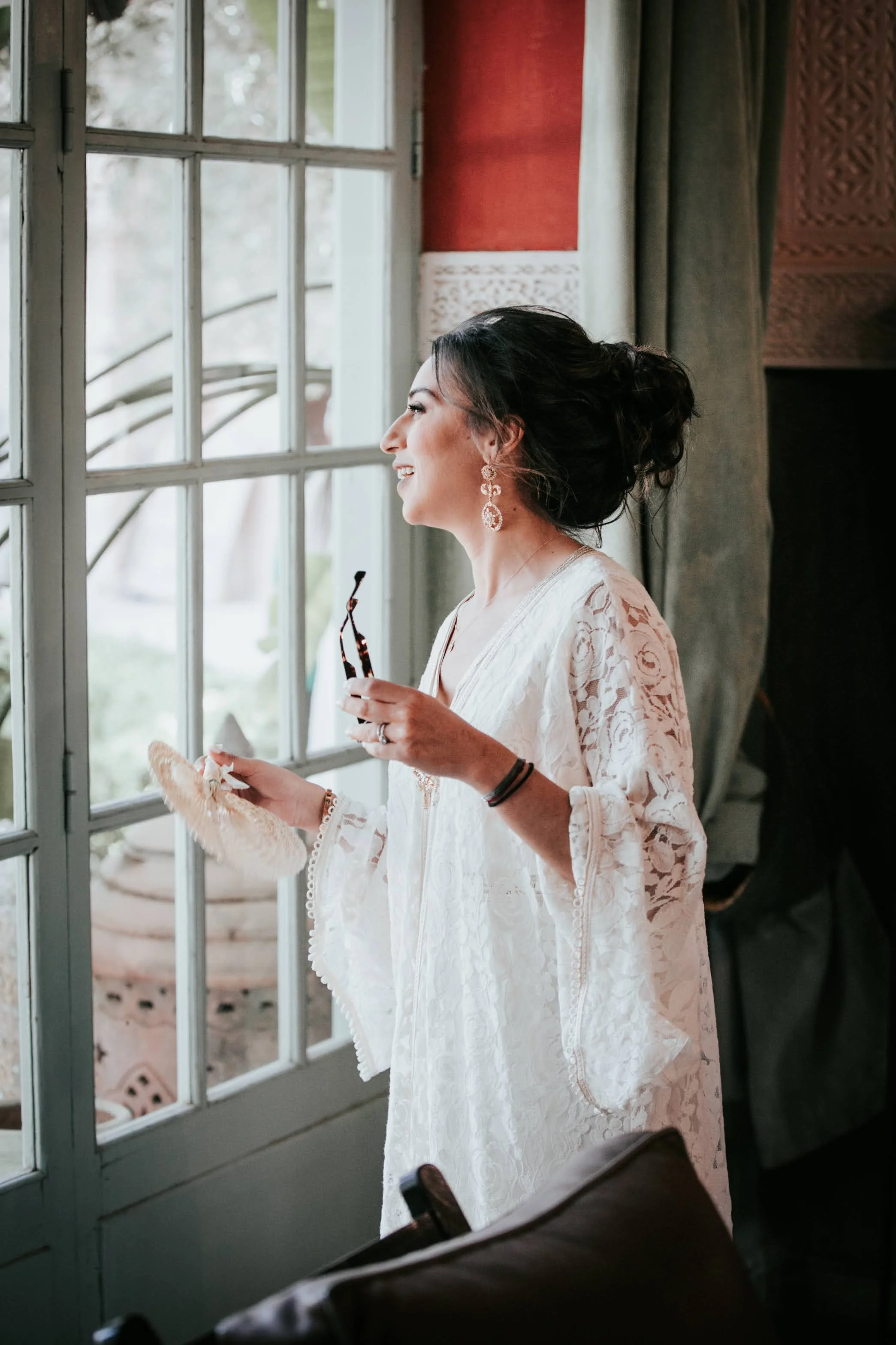 Une femme souriante regardant par une grande fenêtre, portant une robe blanche en dentelle, des boucles d'oreilles pendantes, tenant une paire de lunettes de soleil et un éventail.
