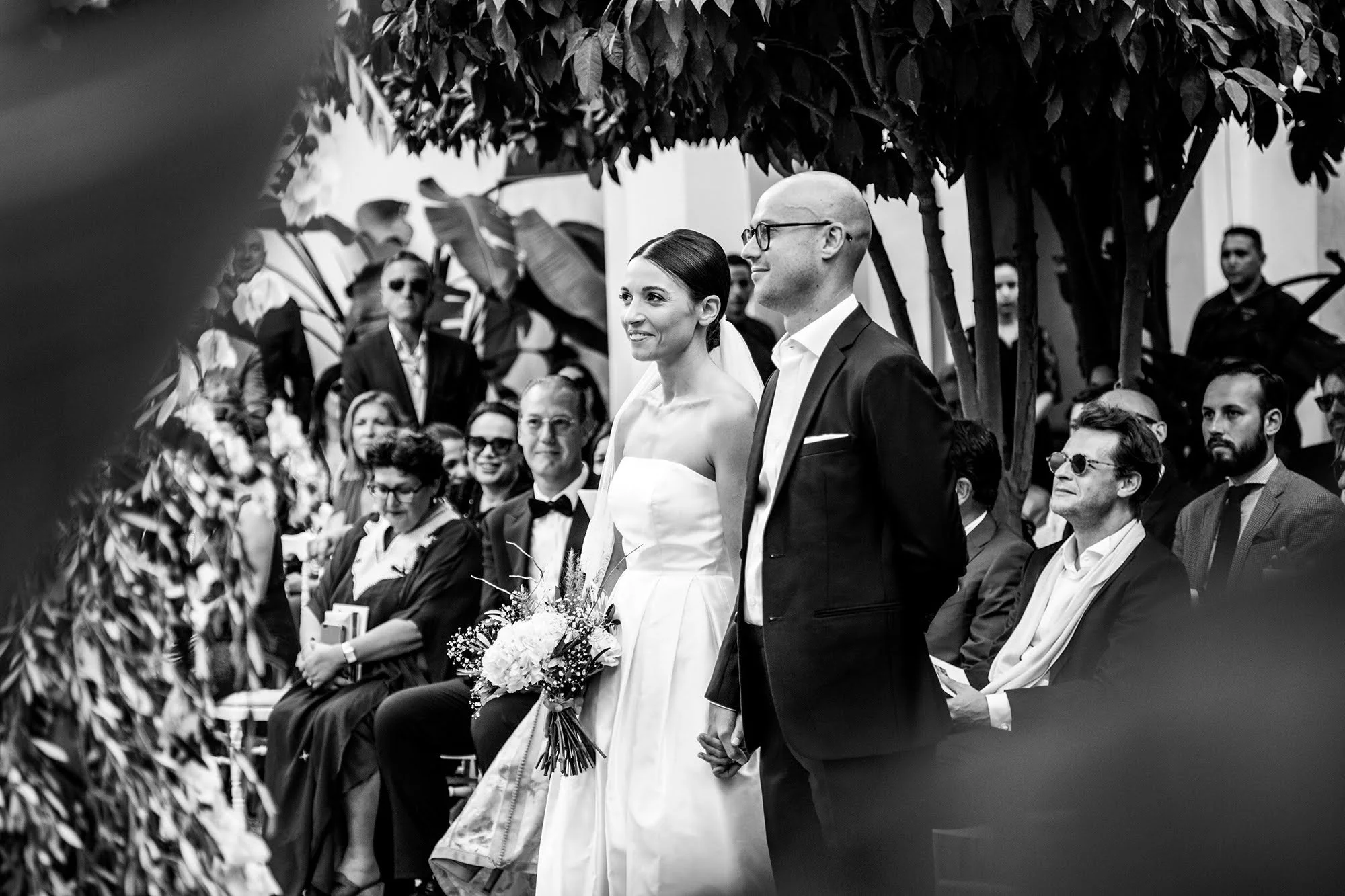 Un couple lors d'un mariage, debout en tenue élégante, entouré d'invités assis et de végétation luxuriante, en noir et blanc.