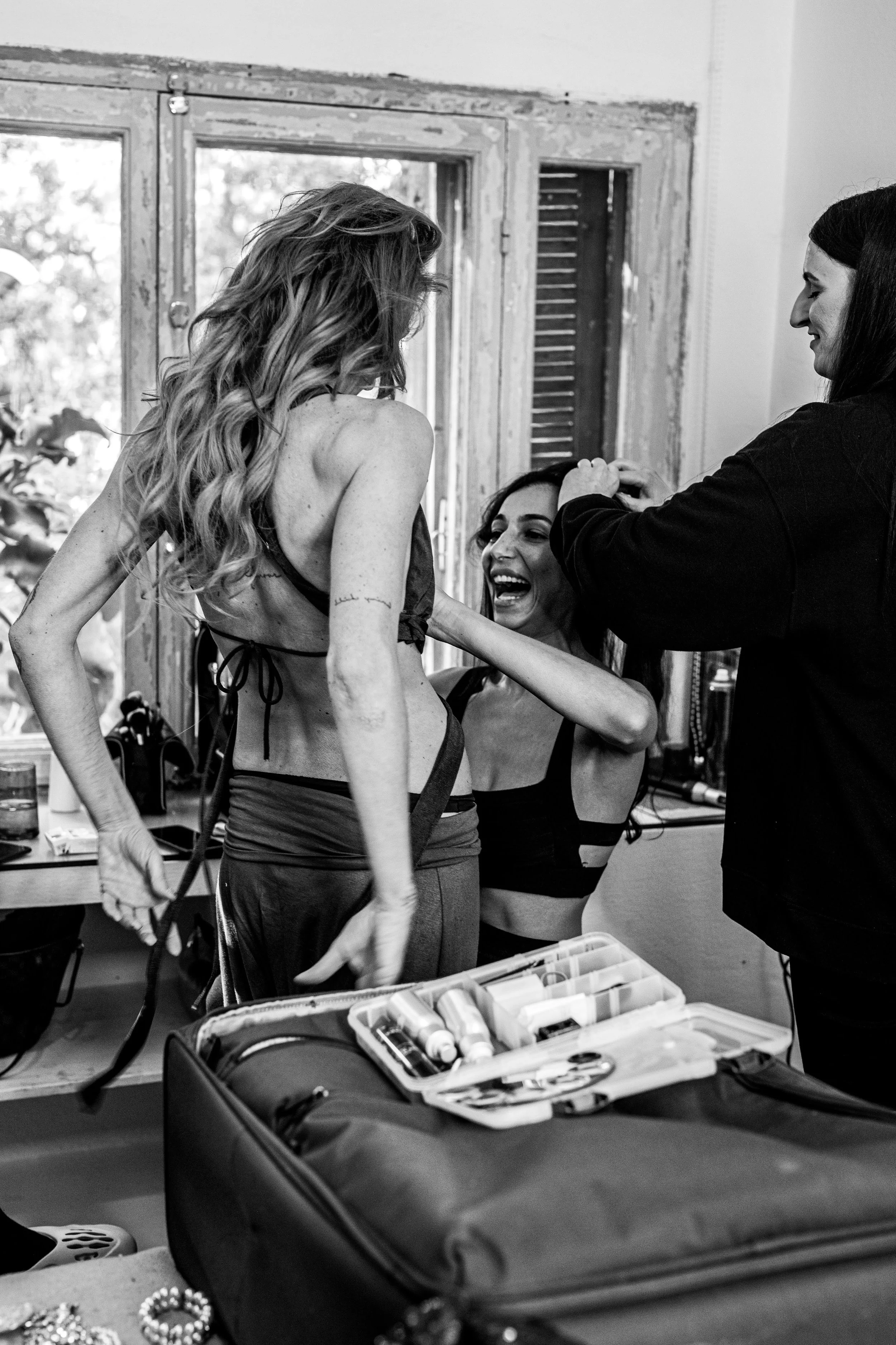 Trois femmes en train de se préparer, l'une d'elles en sous-vêtements, dans une pièce avec une fenêtre en arrière-plan, sous la supervision d'une autre femme.