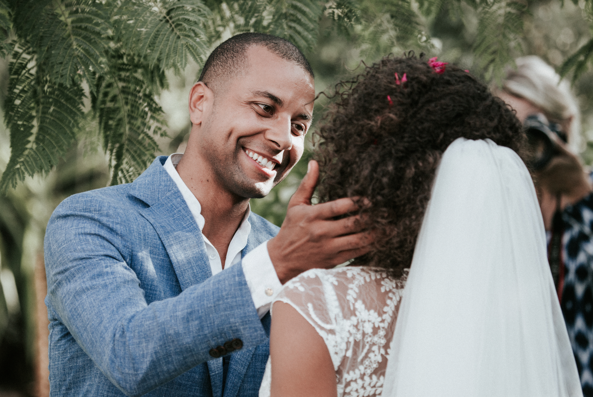 Un homme en costume bleu souriant lors d'une cérémonie de mariage, tenant la tête d'une femme portant un voile blanc.