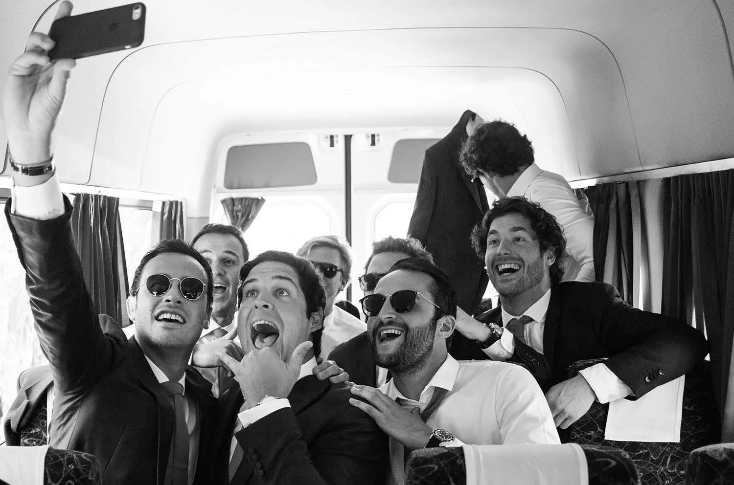 Un groupe de jeunes hommes en costume prennent un selfie dans un bus, souriant et s'amusant.