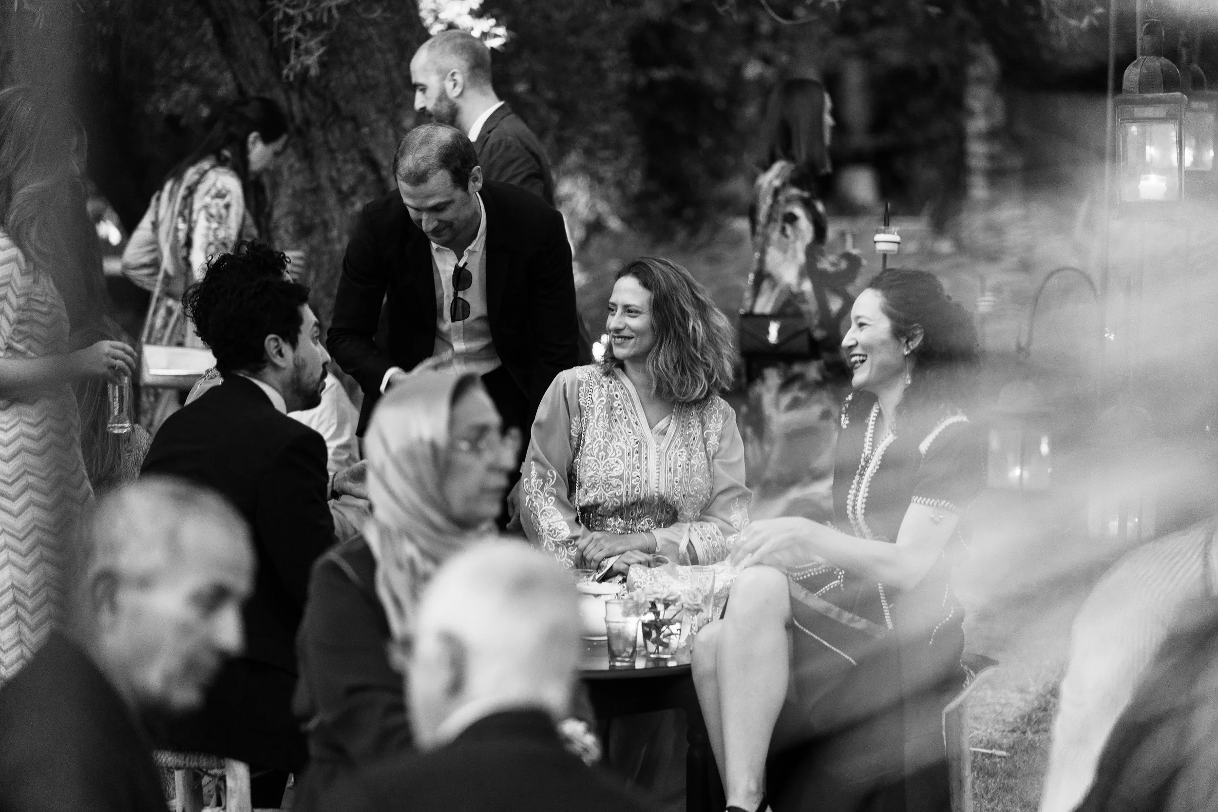 Groupe de personnes lors d'une réunion ou d'une fête en plein air, discutant et souriant, avec des lampes suspendues en arrière-plan, en noir et blanc.