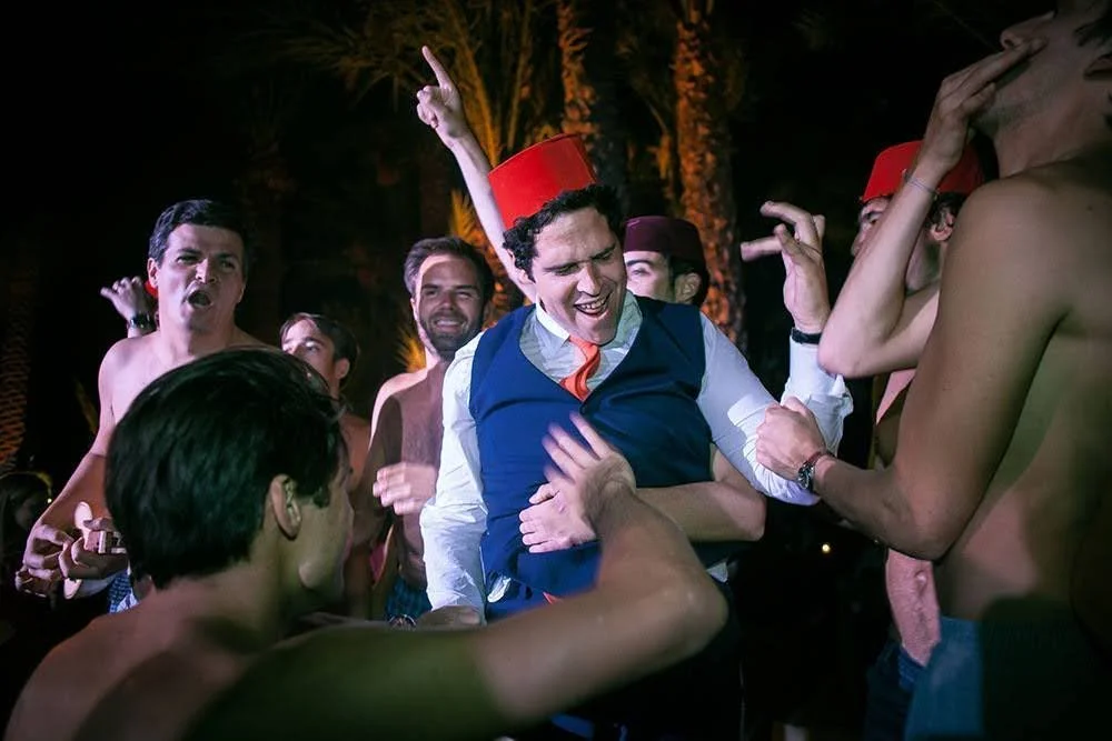 Un homme souriant, habillé en costume, entouré de plusieurs jeunes hommes nus ou en vêtements légers, lors d'une fête en plein air la nuit.