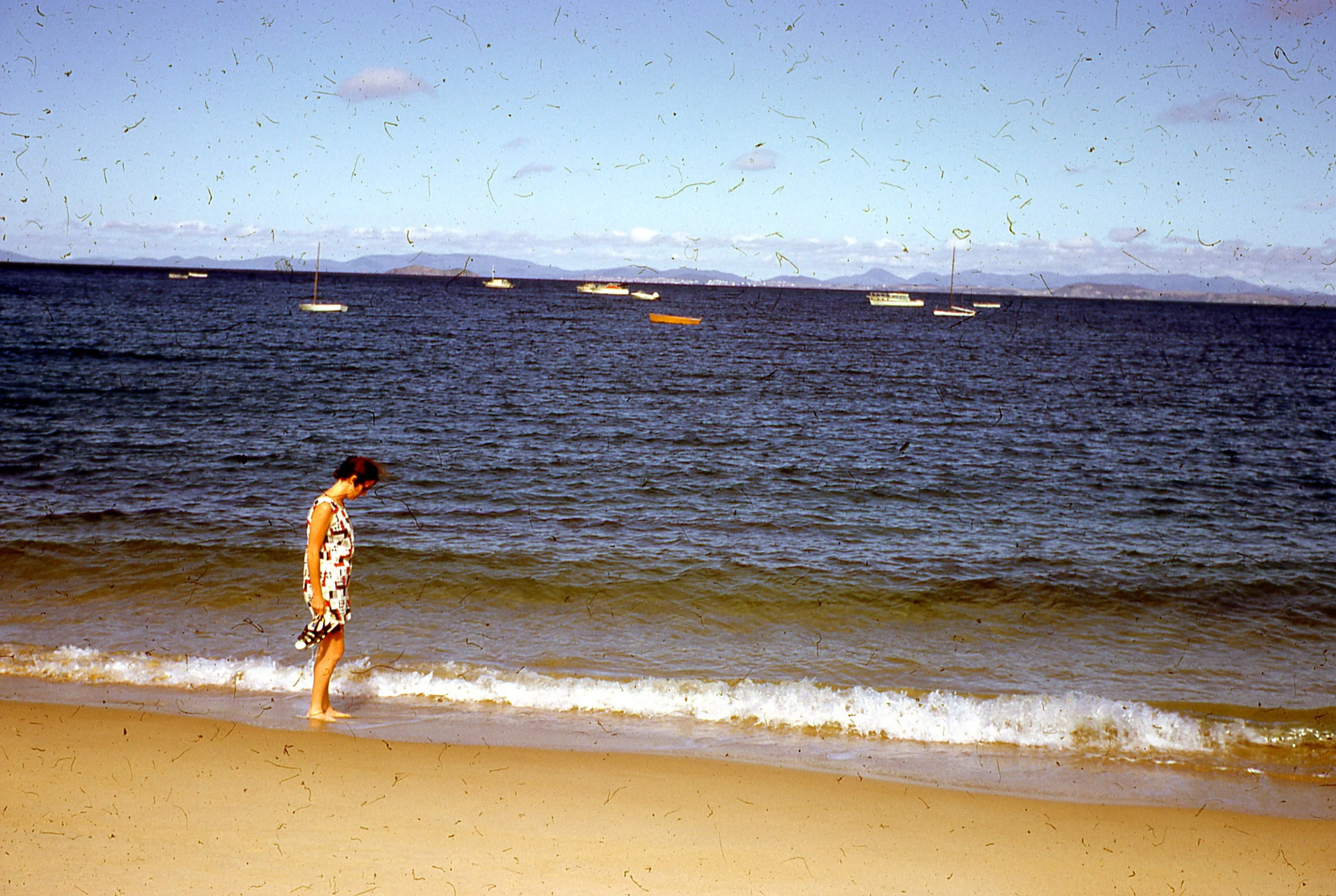 A young girl in a patterned dress standing at the shoreline with small waves touching her feet, looking down at the sand, on a sunny beach with boats floating in the water and mountains in the background.