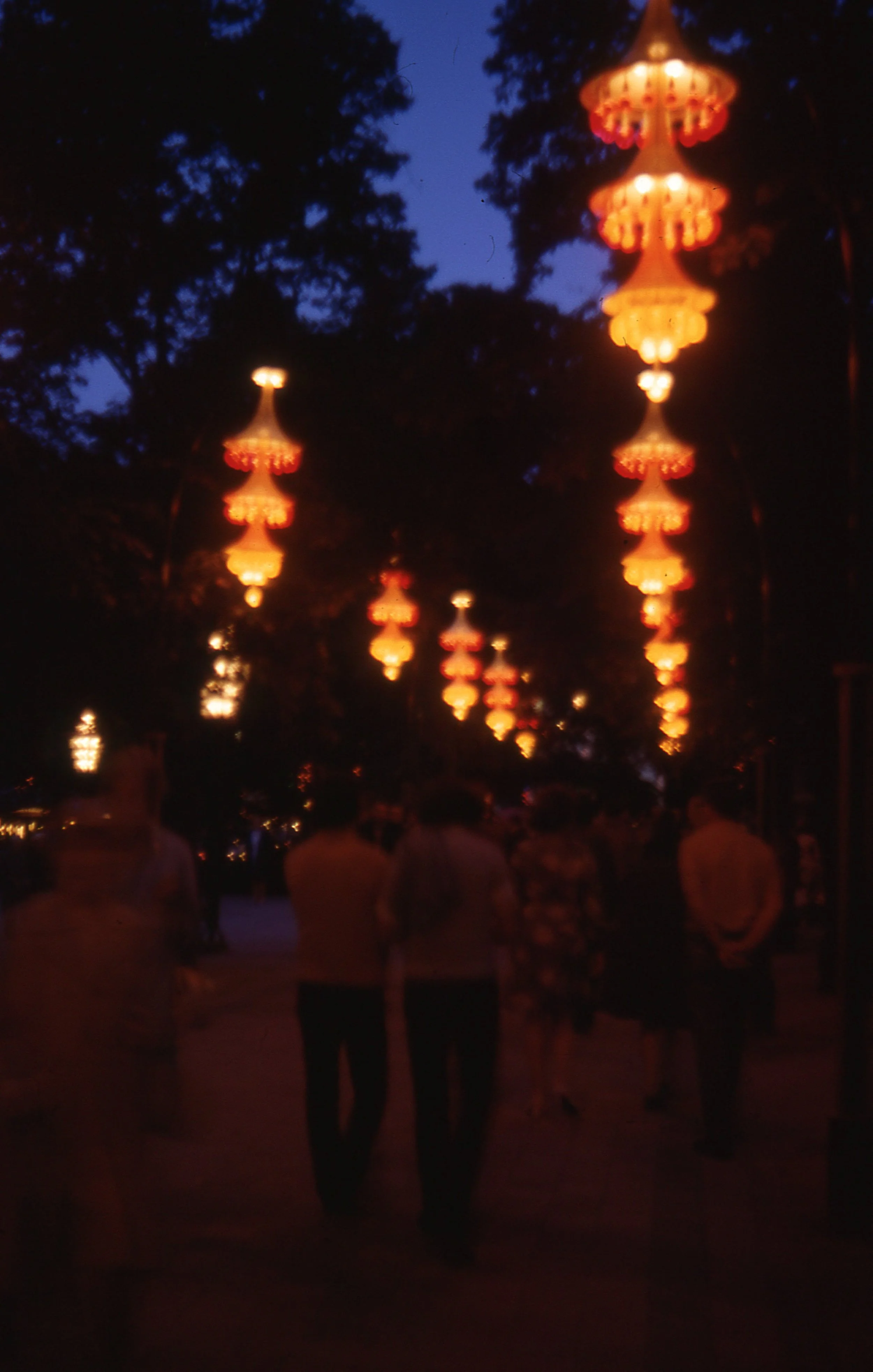 Nighttime scene with hanging lanterns emitting warm glow, trees in the background, and a group of people walking along a path.