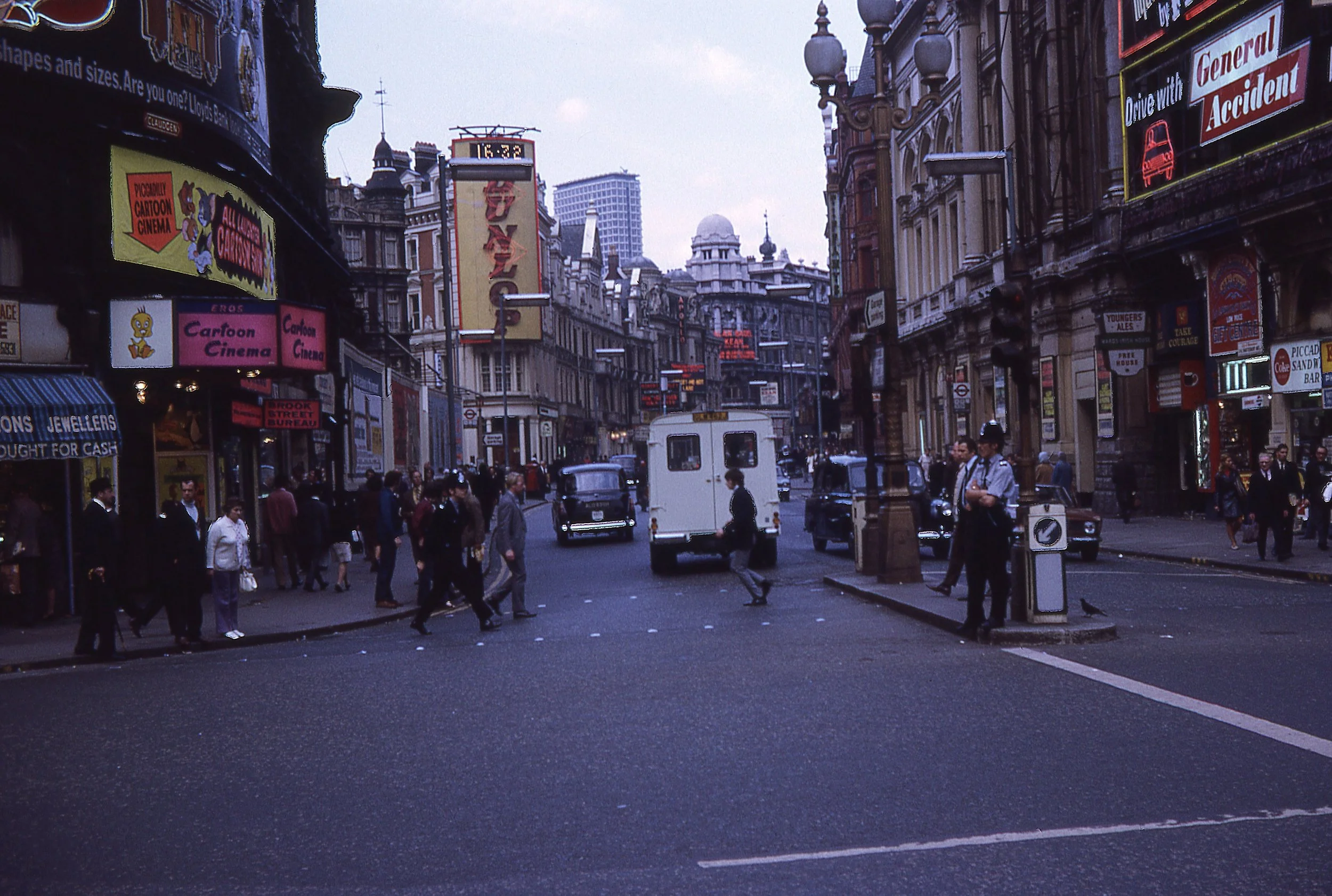 A busy city street scene in London, with pedestrians crossing the street, black cabs, and vintage-style cars, surrounded by tall old buildings adorned with colorful billboards and signs, including a large marquee for a cinema.