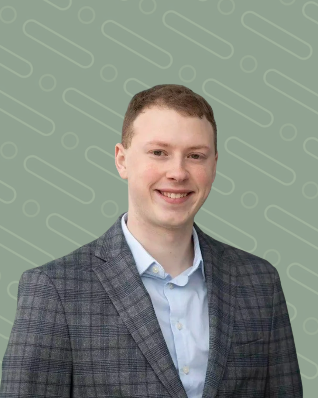 A young man with short brown hair, wearing a light blue collared shirt and a gray plaid blazer, smiling outdoors in front of a modern glass building.