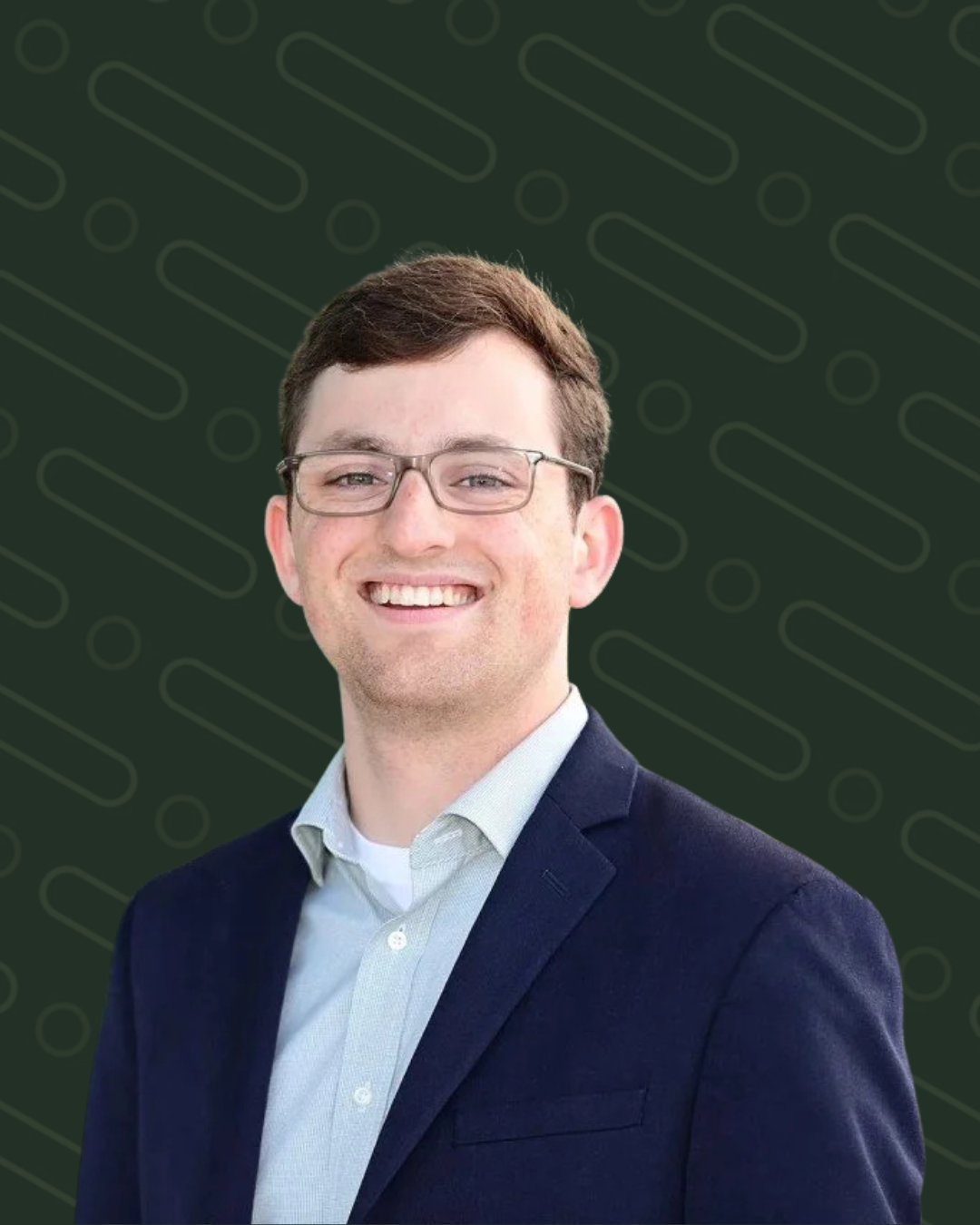 A young man with short brown hair, glasses, and a friendly smile, wearing a dark blazer over a light collared shirt, standing outdoors in front of a modern glass building.
