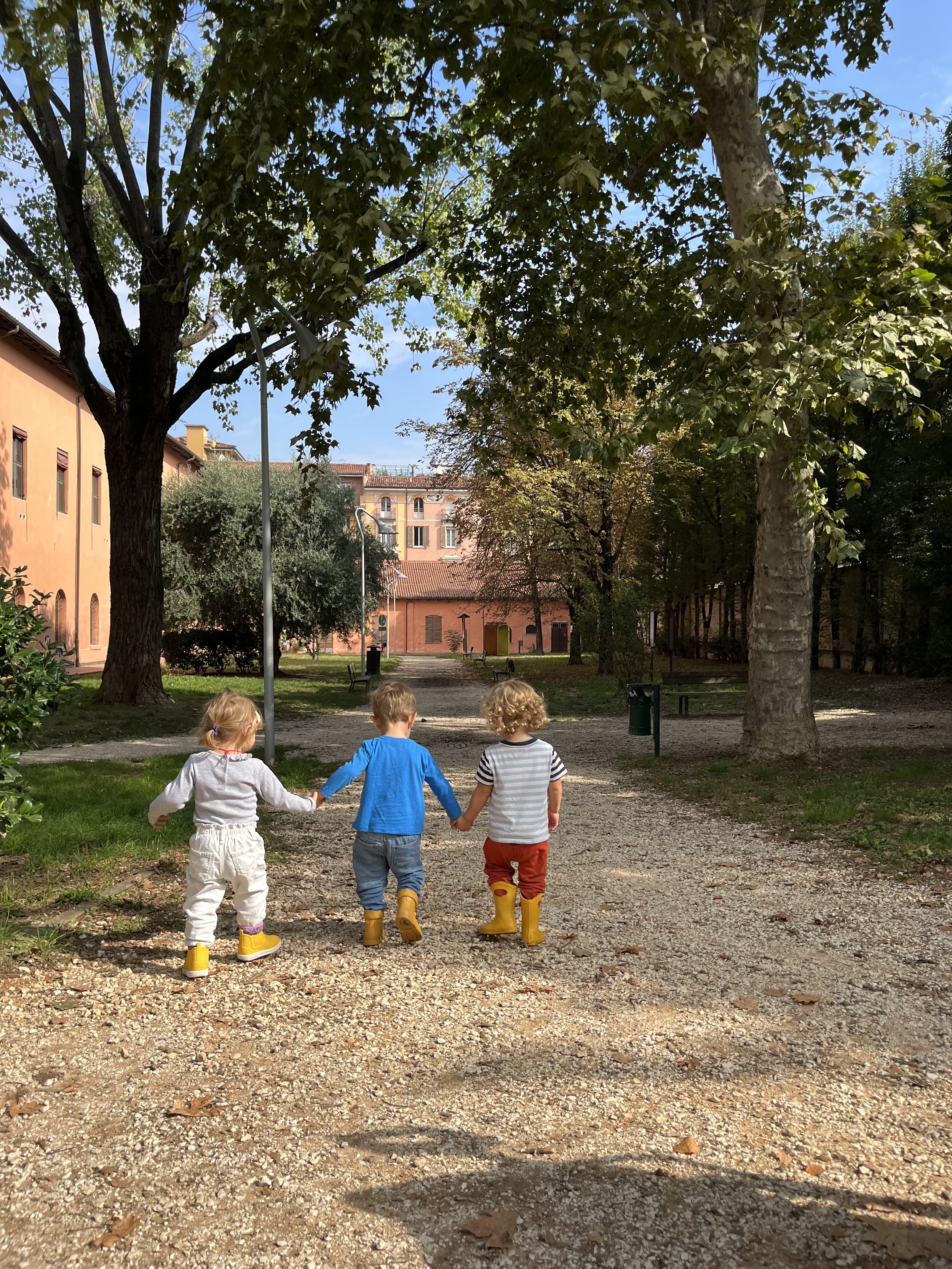 Tre bambini con stivali di gomma che si tengono per mano mentre camminano lungo un sentiero in un parco alberato con edifici in pietra rosa alle spalle, sotto un cielo blu con alcune nuvole.