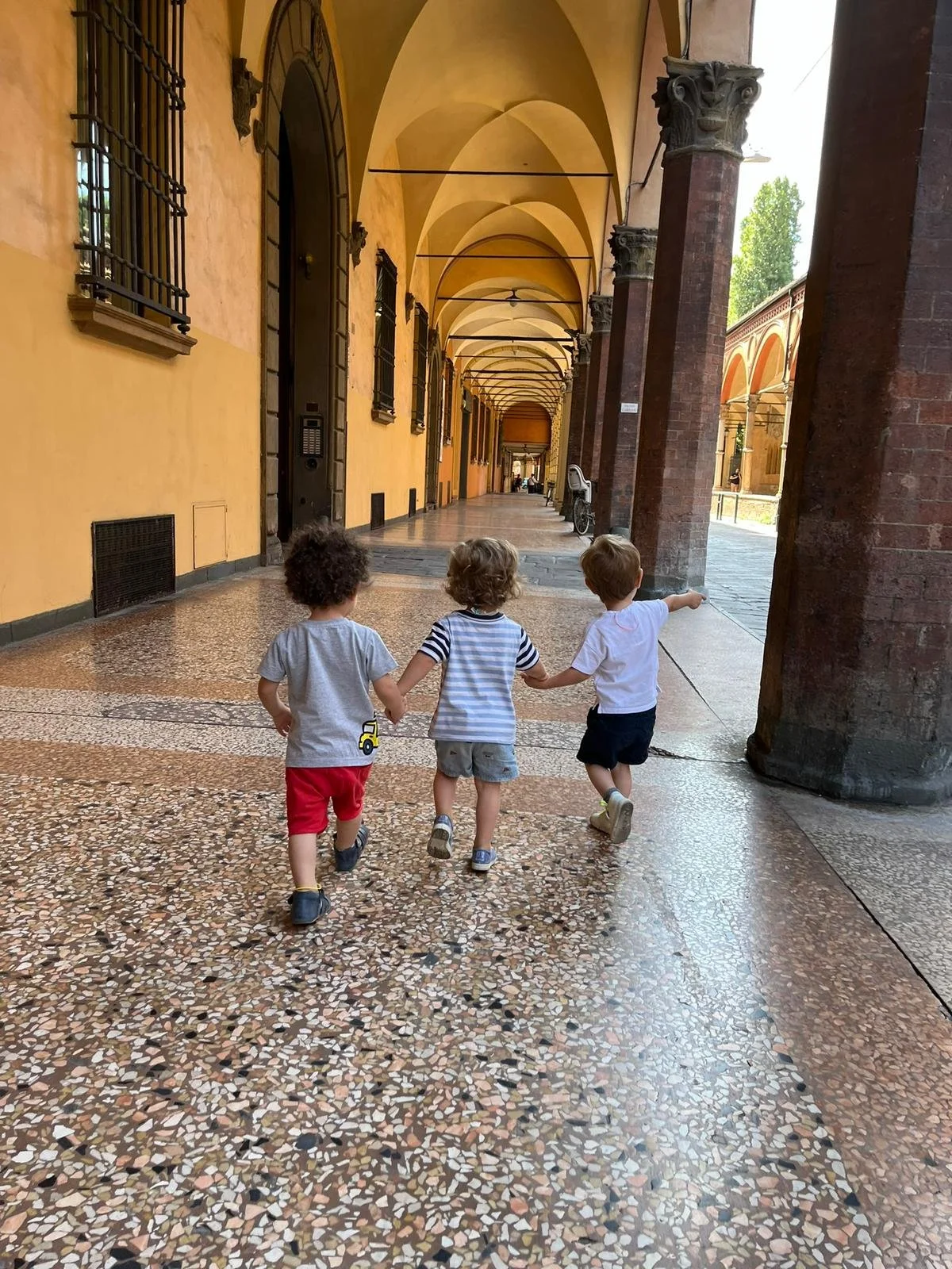 Tre bambini che camminano mano nella mano lungo un portico storico con archi e colonne in pietra, con pavimento in mosaico, in un ambiente europeo all'aperto.
