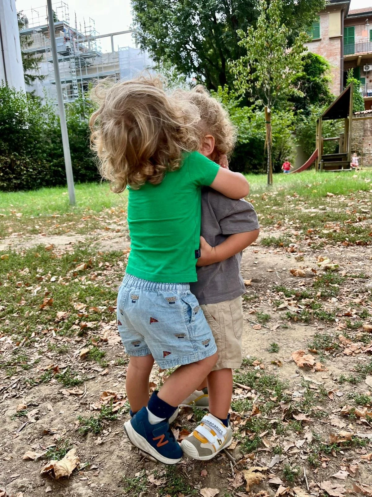 Due bambini abbracciati nel parco, uno con capelli ricci biondi e maglietta verde, l'altro con capelli più corti e maglietta grigia, in un'area con alberi, foglie sul terreno, giochi e strutture di costruzione sullo sfondo.