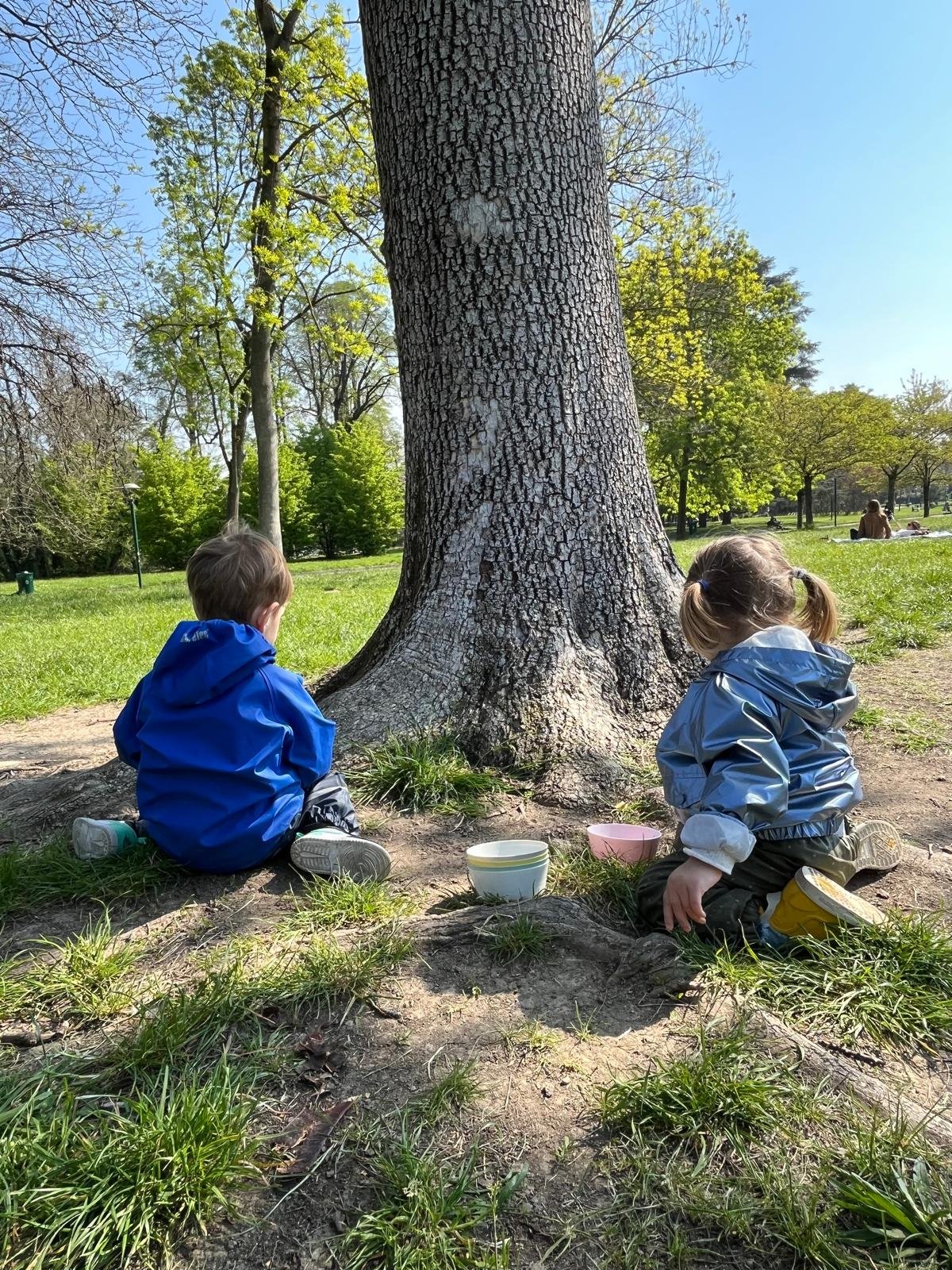 Due bambini siedono sull'erba vicino a un grande albero in un parco, con ciotole colorate davanti a loro, in una giornata di sole con cielo limpido.