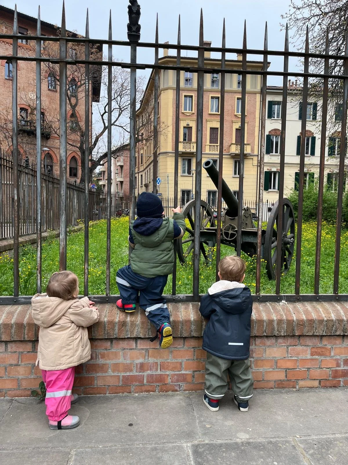 Tre bambini osservano un cannone antico dietro una recinzione in ferro, con edifici residenziali sullo sfondo.