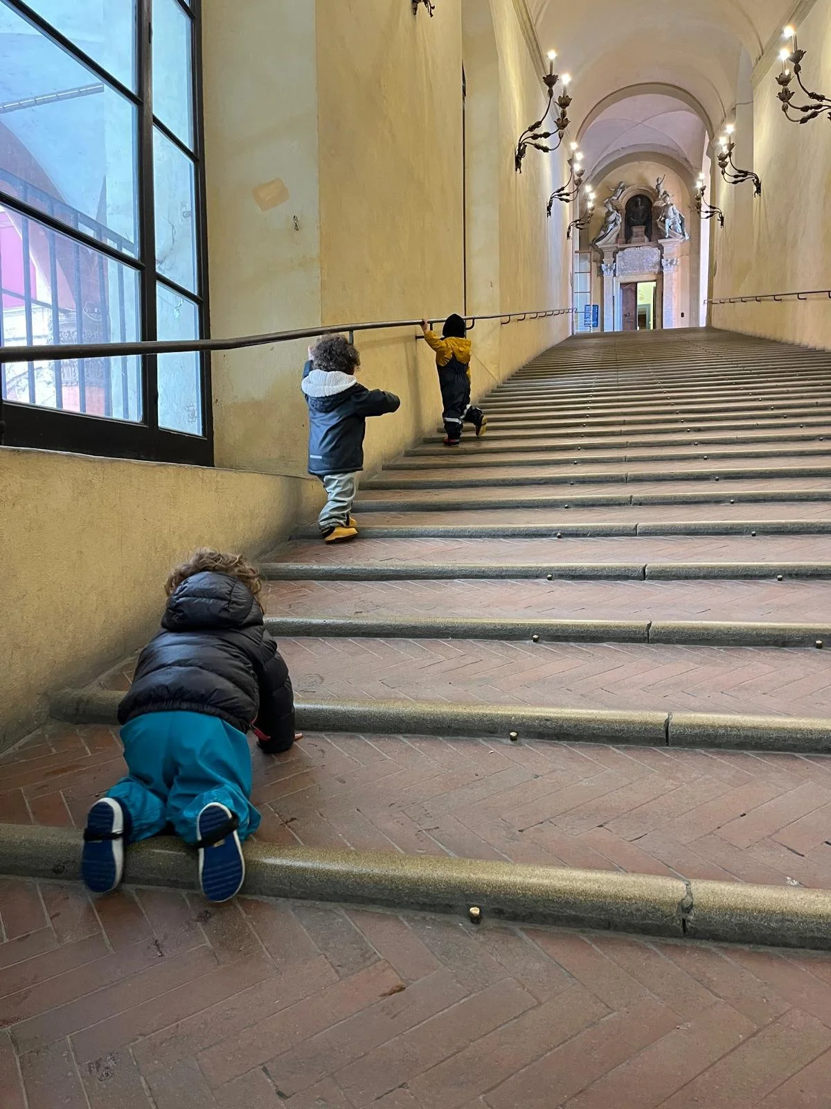 Tre bambini piccoli che salgono una grande scalinata in un edificio storico con pareti gialle e lampadari decorativi.