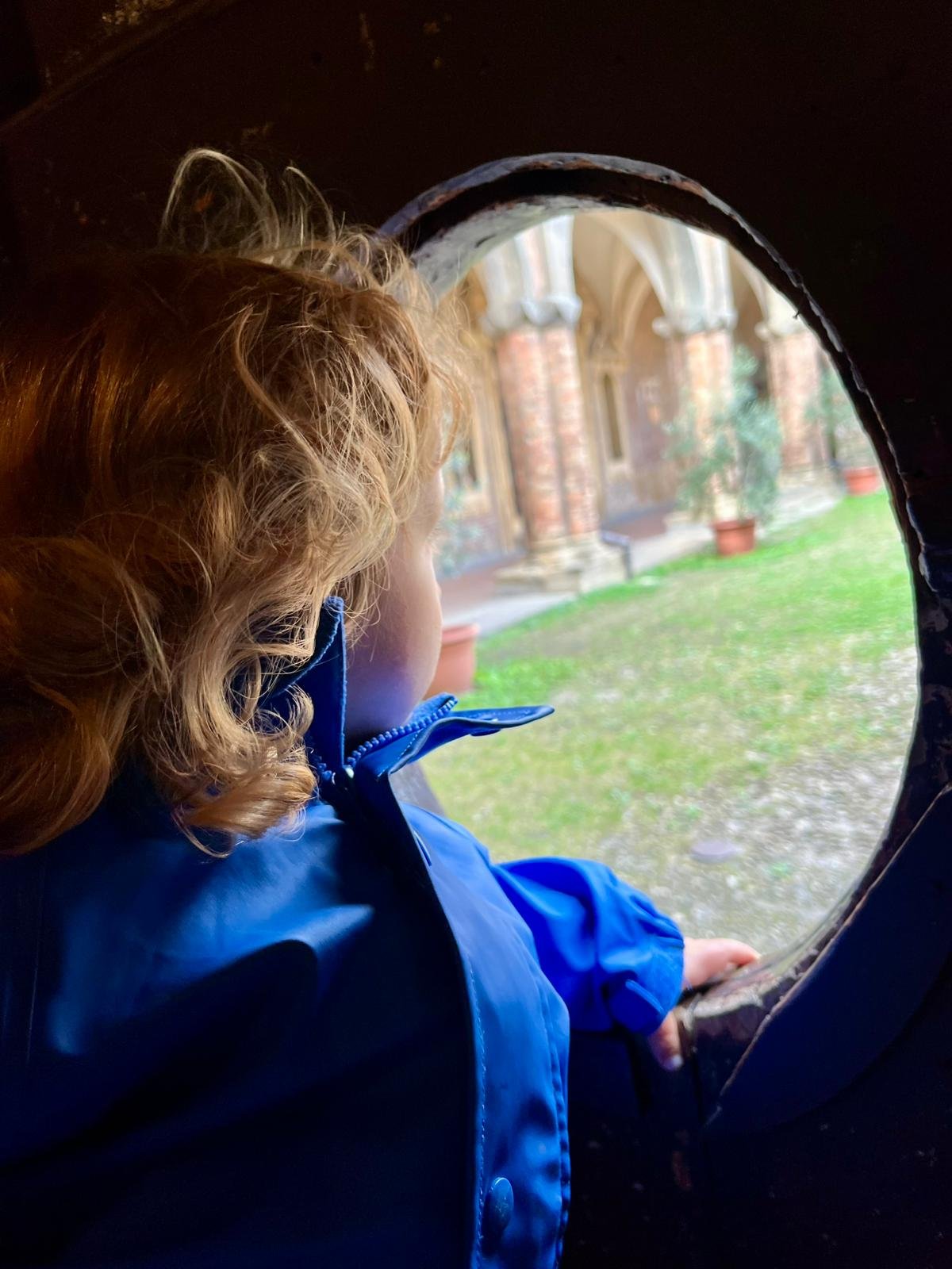 Bambino con capelli ricci perlacei e giacca blu, che guarda fuori da un fornice in una struttura di legno, con un cortile con erba, piante e un edificio storico in pietra e mattoni in fondo.