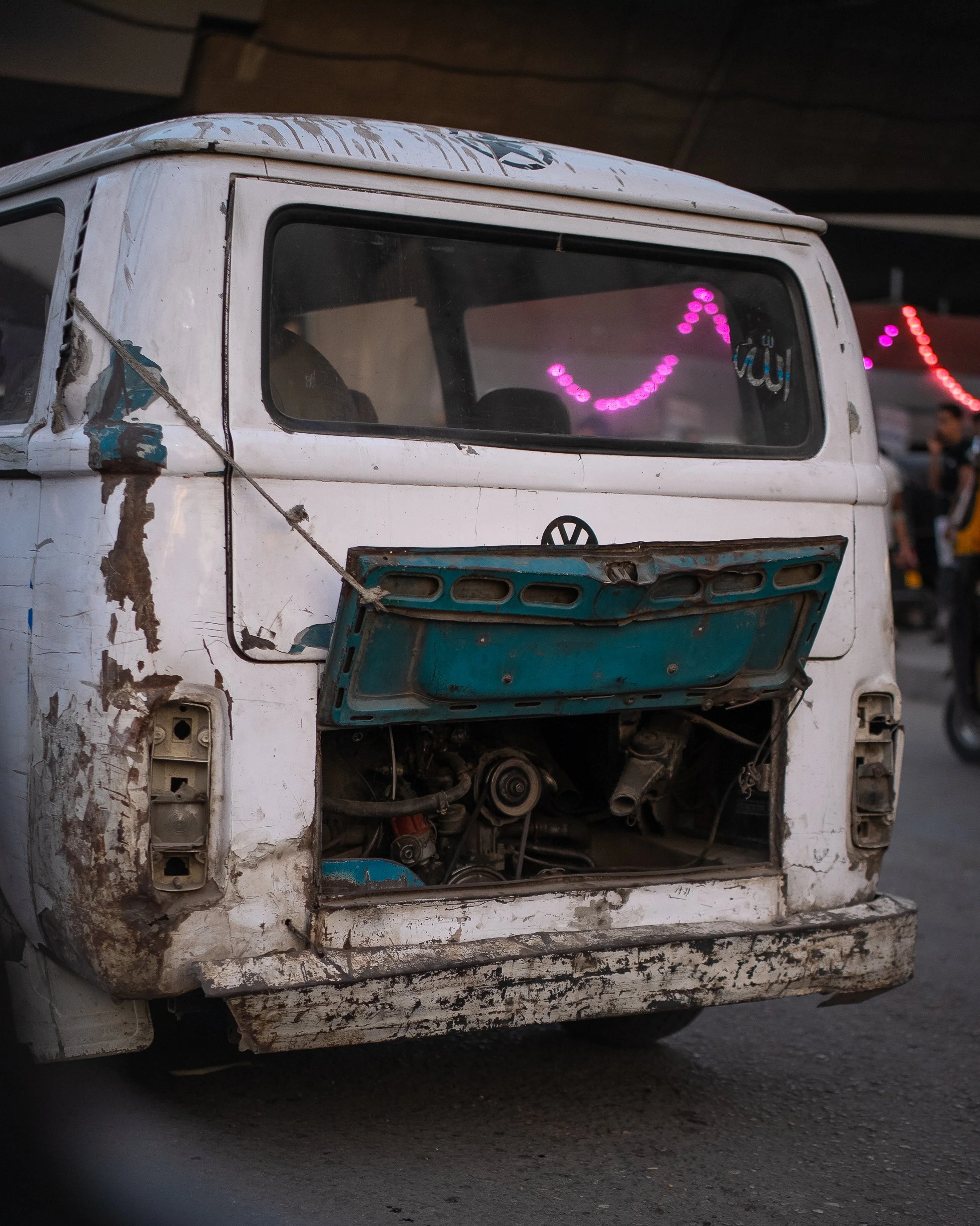 An old, distressed Volkswagen van with missing front panel and exposed engine, showing signs of rust and peeling paint, parked on a street.